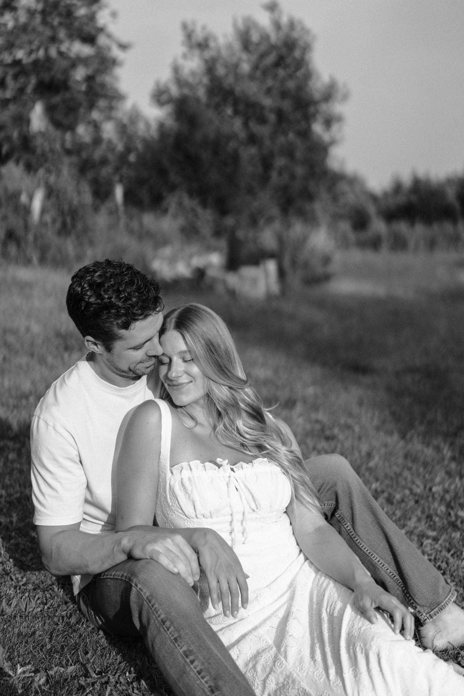 A romantic black and white portrait of a couple relaxing together on grassy ground with trees in the background.
