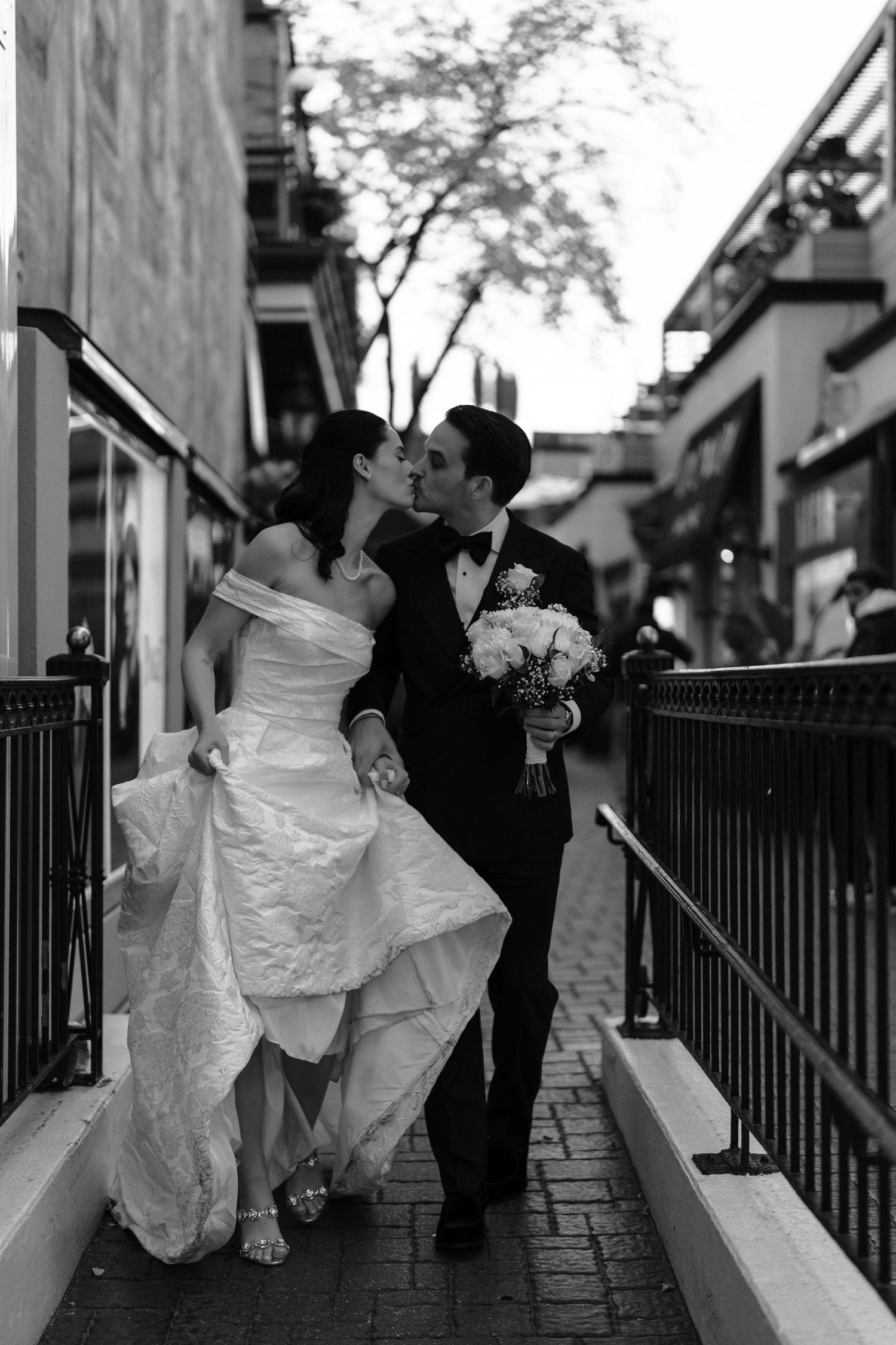 A romantic black and white wedding photo showing a couple embracing on a brick walkway with urban architecture.