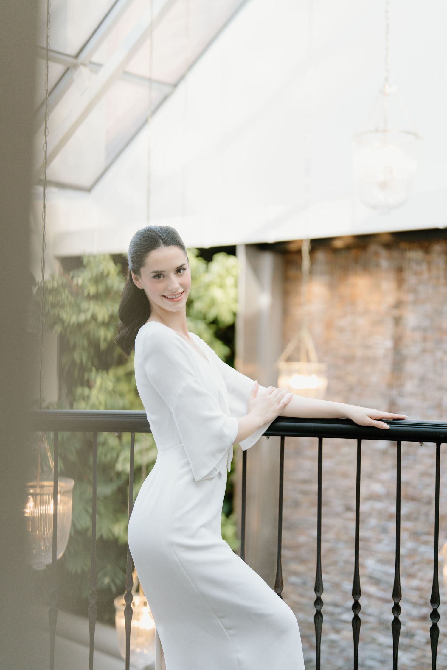 A person in a white dress poses gracefully on a balcony with natural lighting and architectural details.