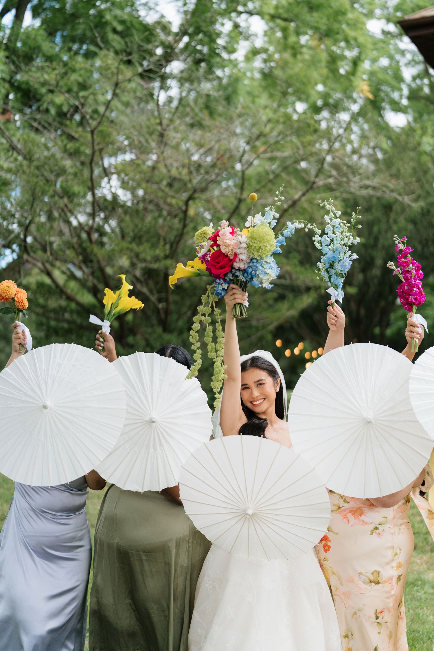 Wedding party holds white paper parasols and colorful bouquets while standing in a row outdoors.
