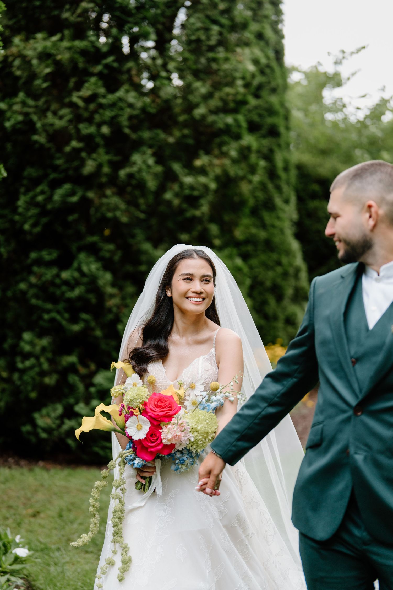 A joyful wedding couple shares candid moments in a garden setting with colorful bouquet and green suit attire.