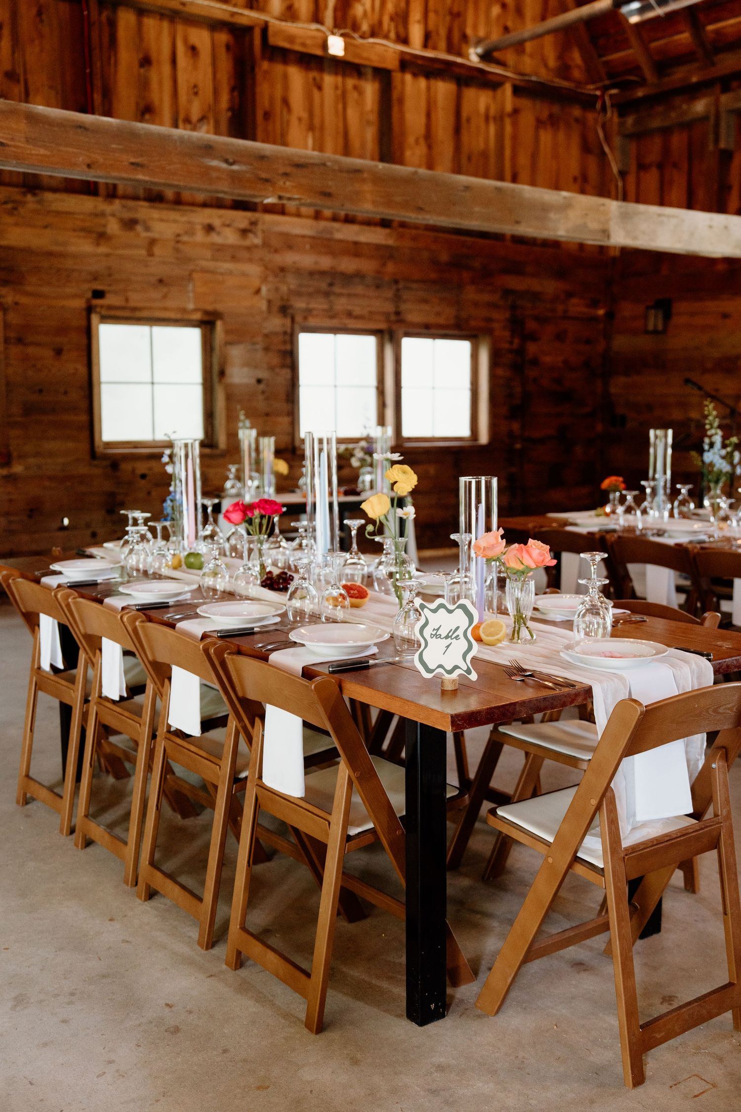 Rustic barn wedding reception setup with wooden chairs and tables arranged in rows under exposed beams.