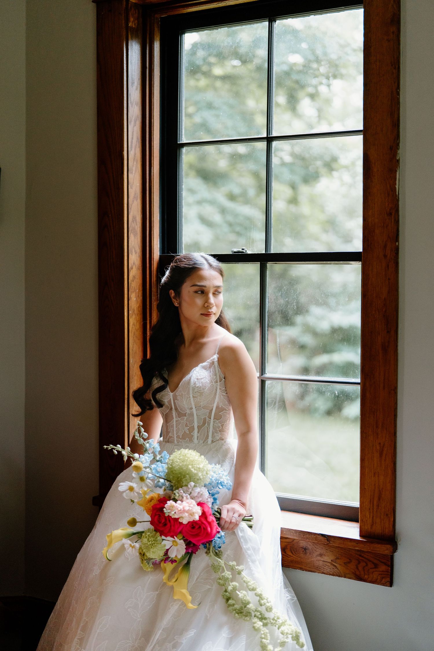 A bride in a white wedding gown stands by a window holding a colorful bouquet of flowers in a rustic interior.