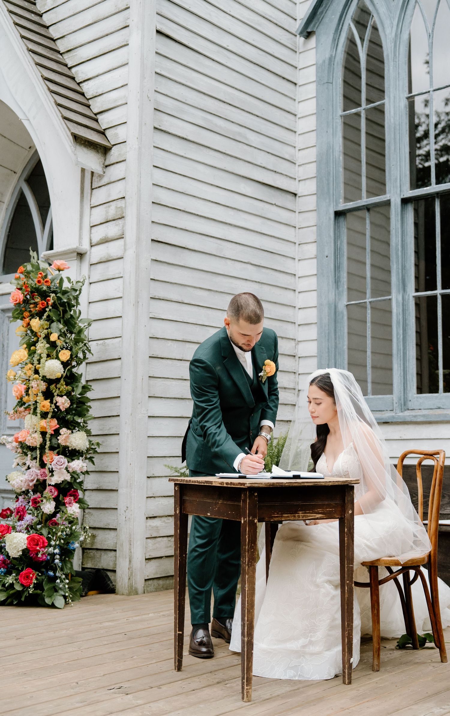 A couple signs their marriage license at a rustic table outside a white chapel with floral decorations.