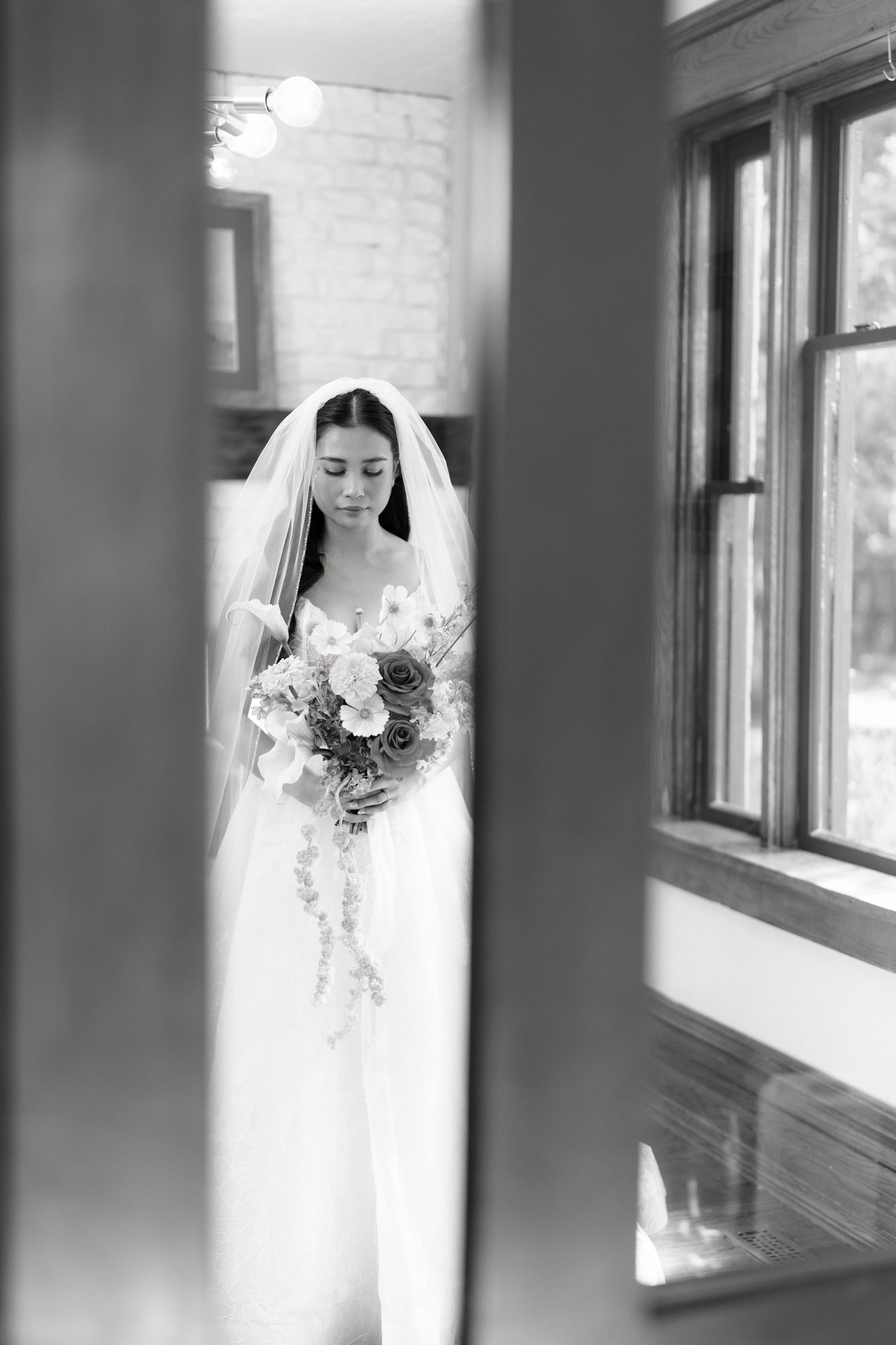A bride in a white wedding gown and veil stands by a window holding a cascading bouquet in this black and white photo.