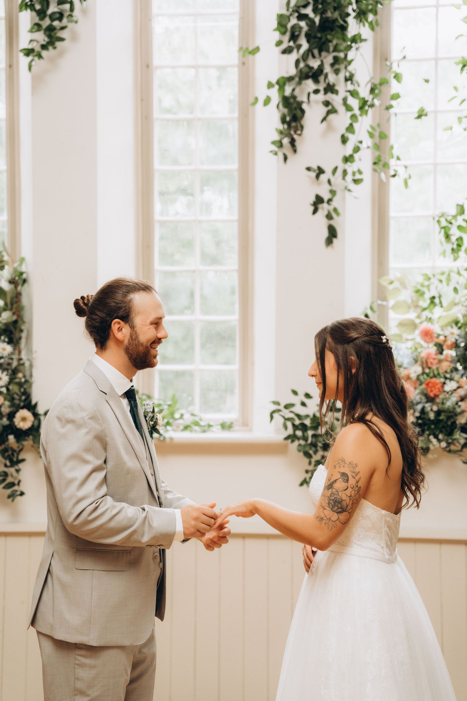 A couple holds hands during their wedding ceremony in a light-filled room adorned with trailing vines and flowers.