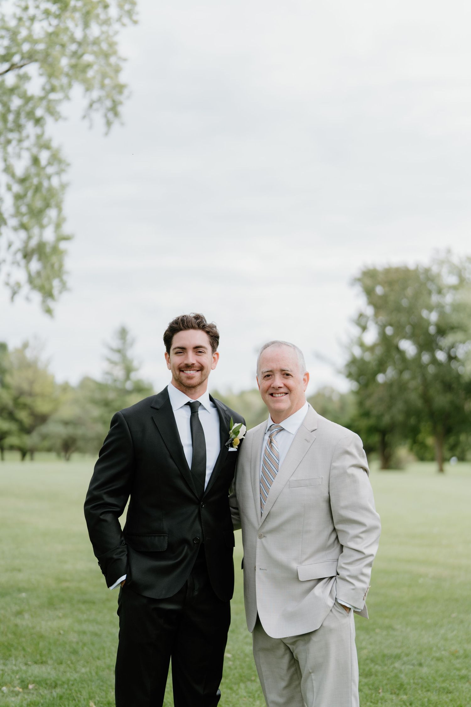 Two men in suits stand together smiling on a grassy lawn with trees in the background.