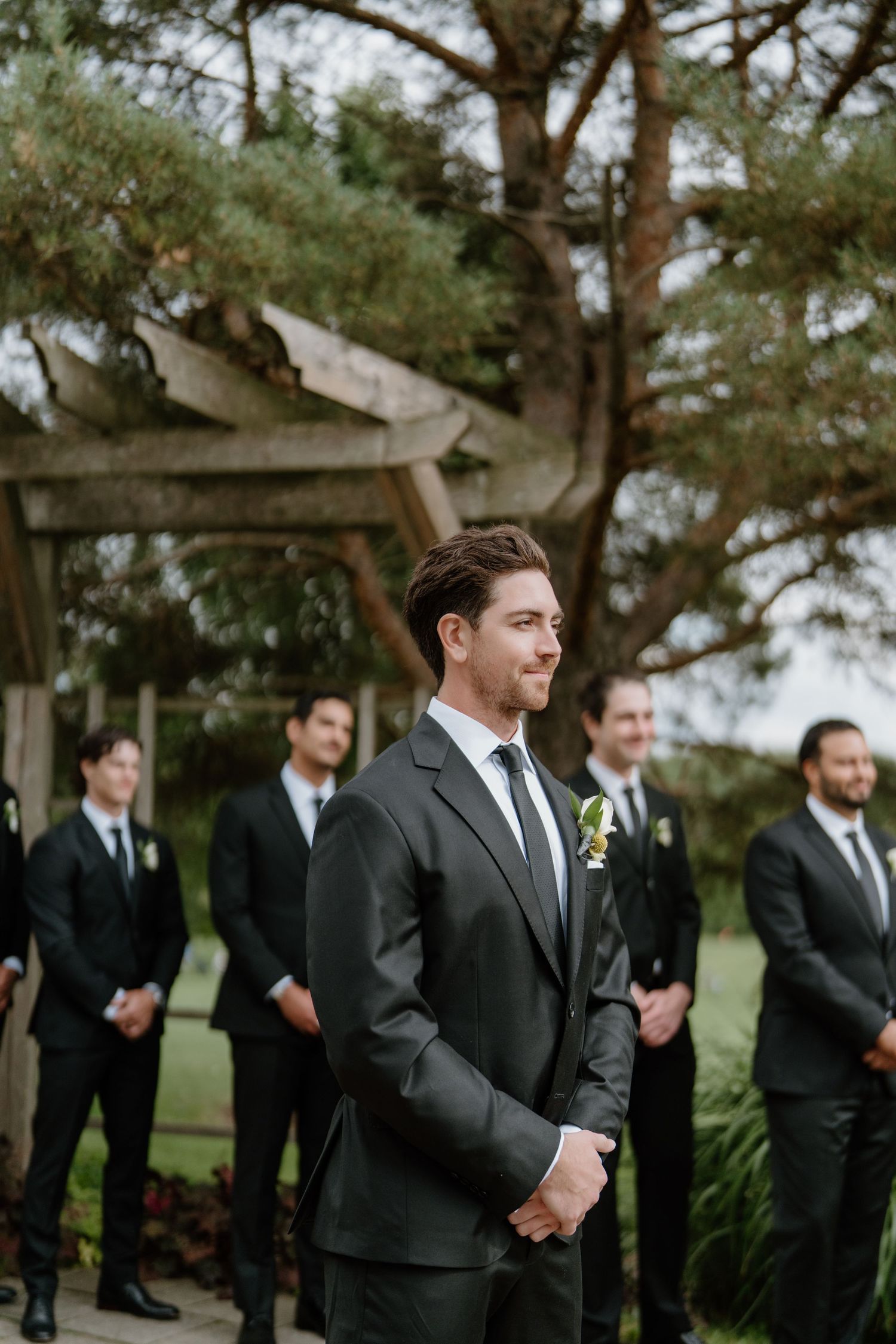 Groomsmen in black suits share a candid moment while standing under trees at an outdoor wedding venue.