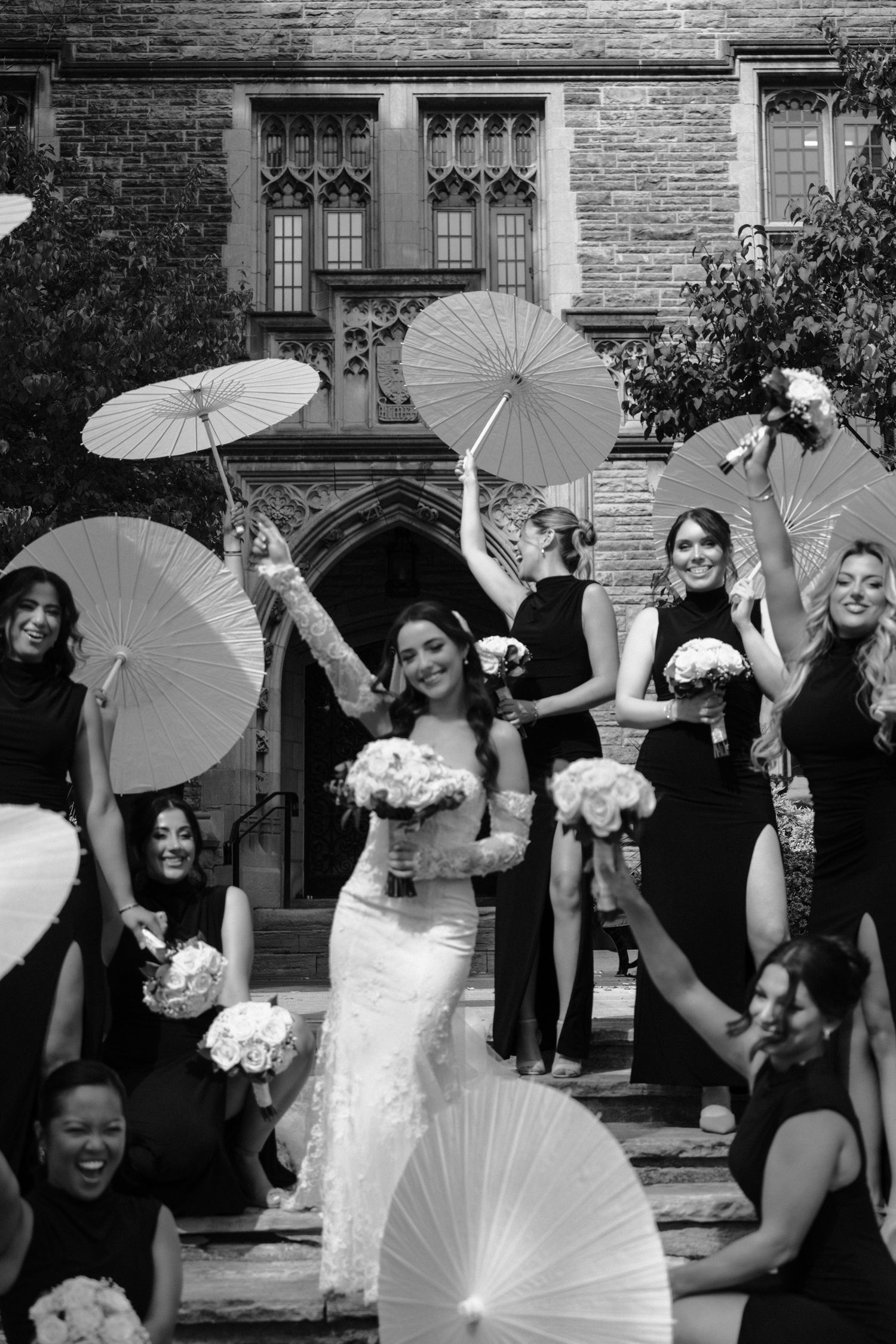 Wedding party poses with white parasols in front of stone building during outdoor celebration.