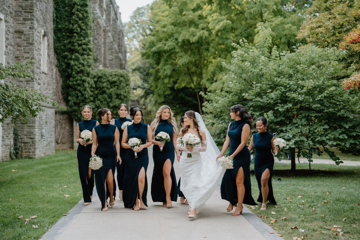 Wedding party in black dresses and white gown pose together in a lush garden setting with stone architecture.