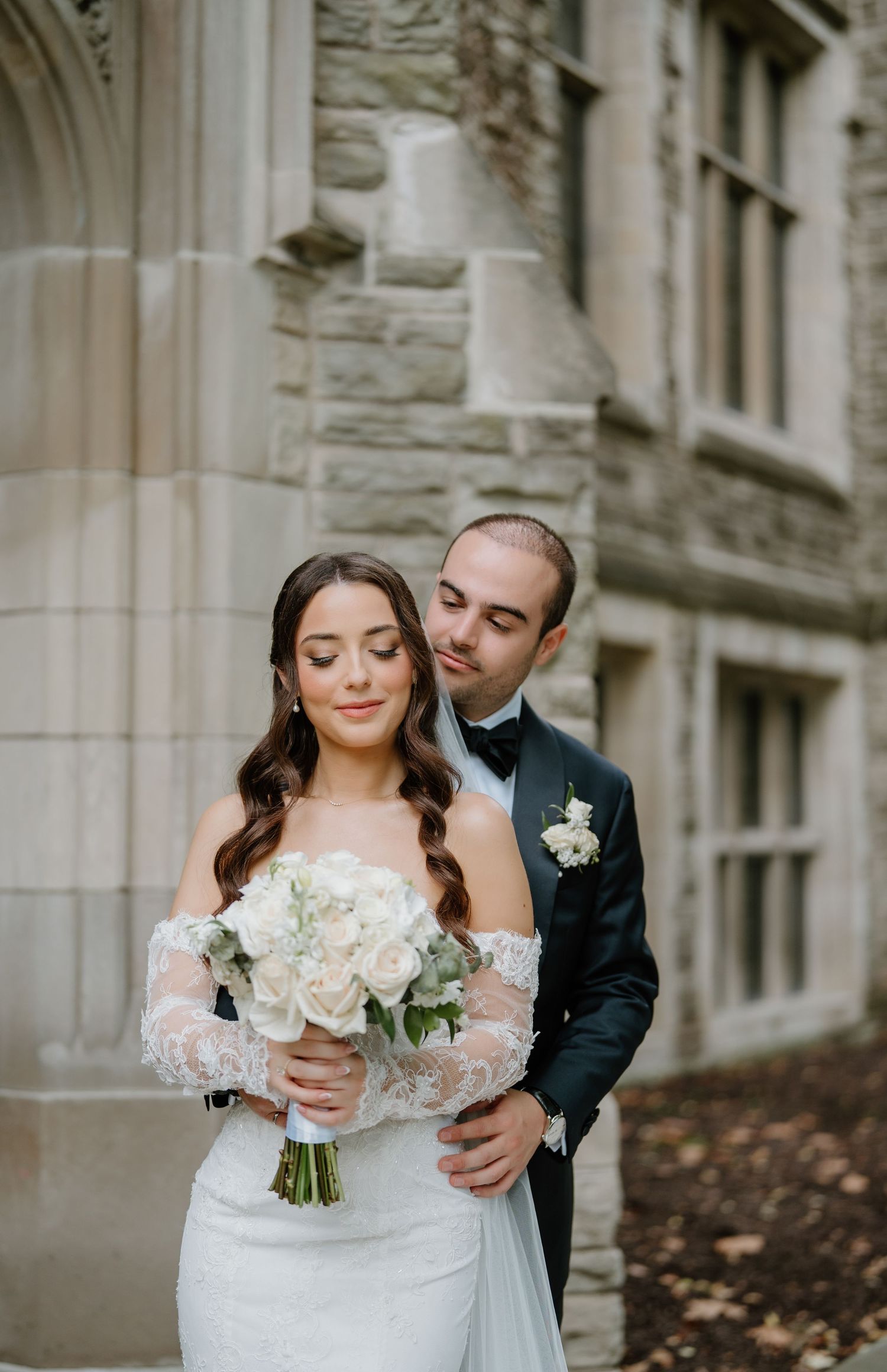 A romantic wedding moment captured in front of a stone building with white floral bouquet and elegant attire.