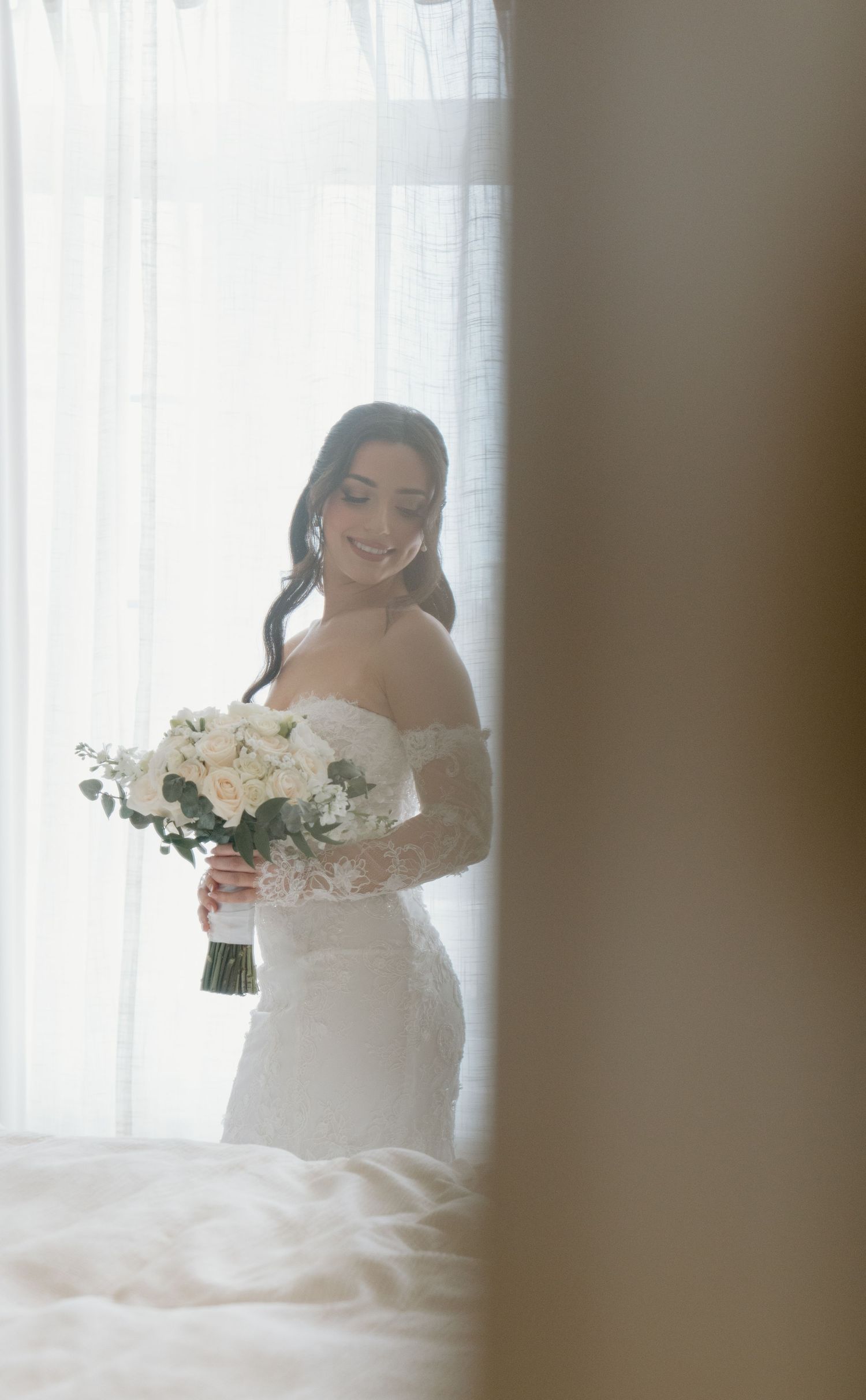 A bride in a white off-shoulder wedding gown holds a bouquet of white flowers while standing by a window.