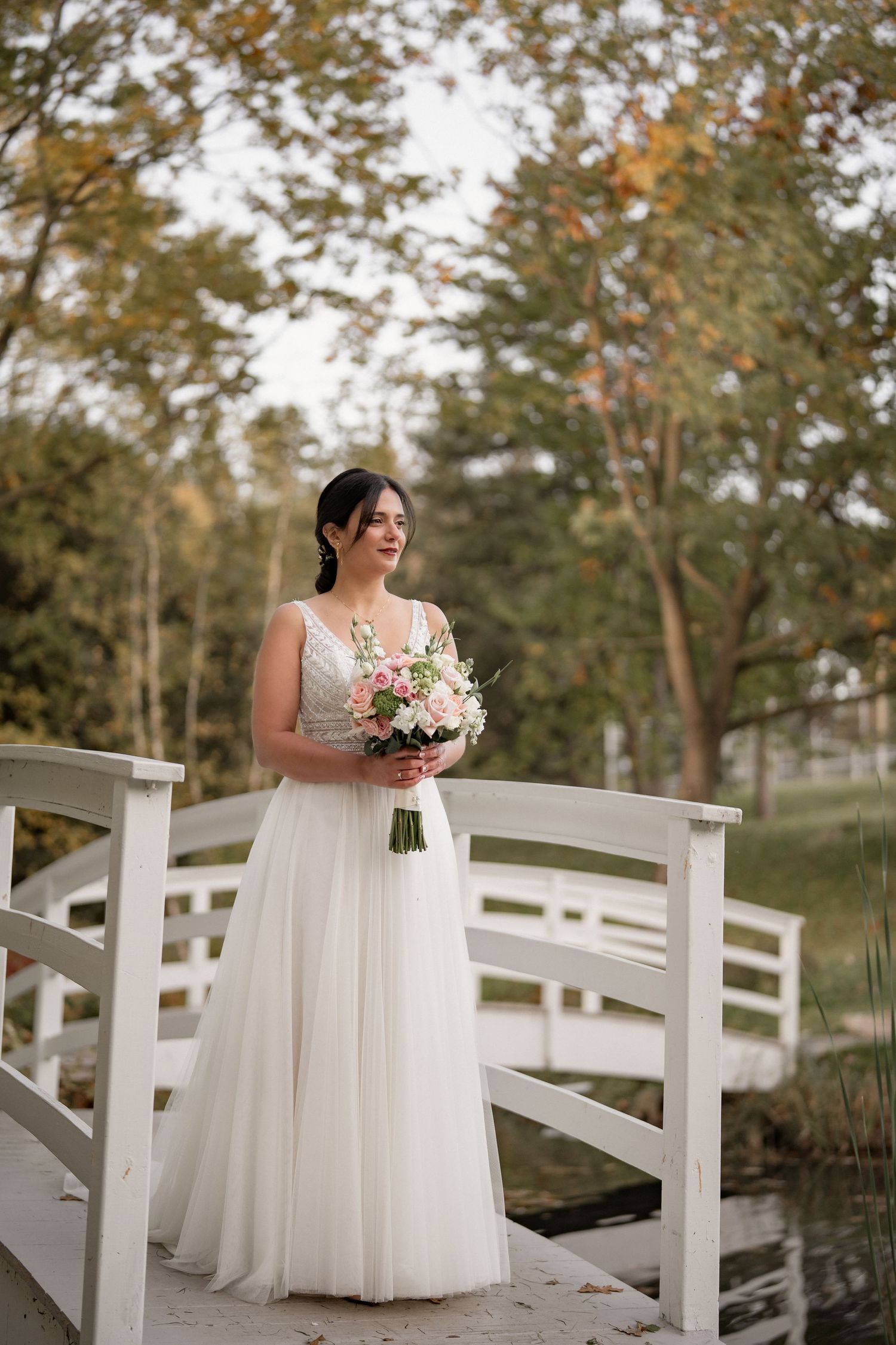 Elegant wedding portrait on a white bridge with bride holding a pastel bouquet among fall foliage.