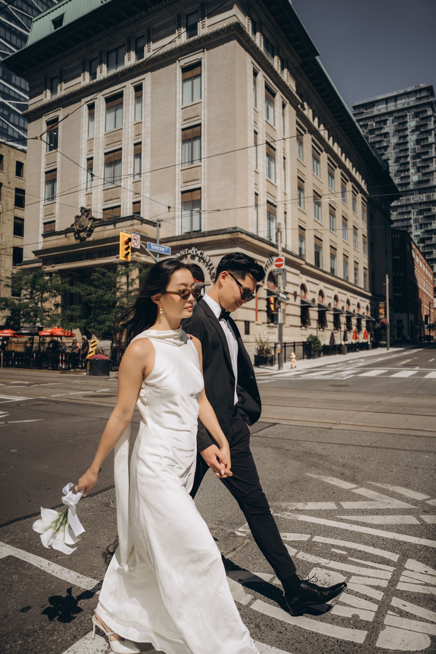 A bride and groom walk across an urban crosswalk together near classic stone buildings in a cinematic sequence.