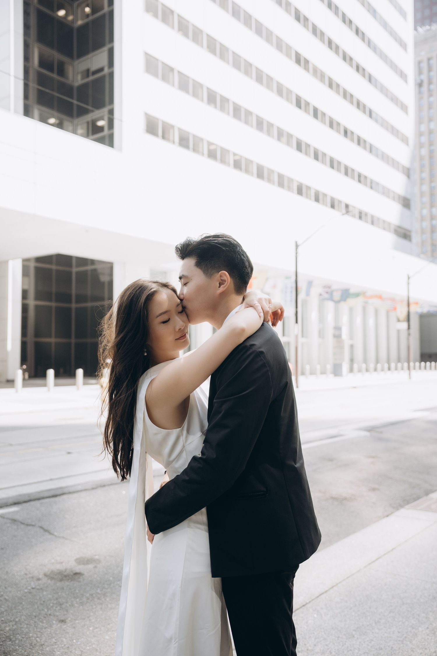 A romantic couple embraces on an urban street with modern white buildings in the background.