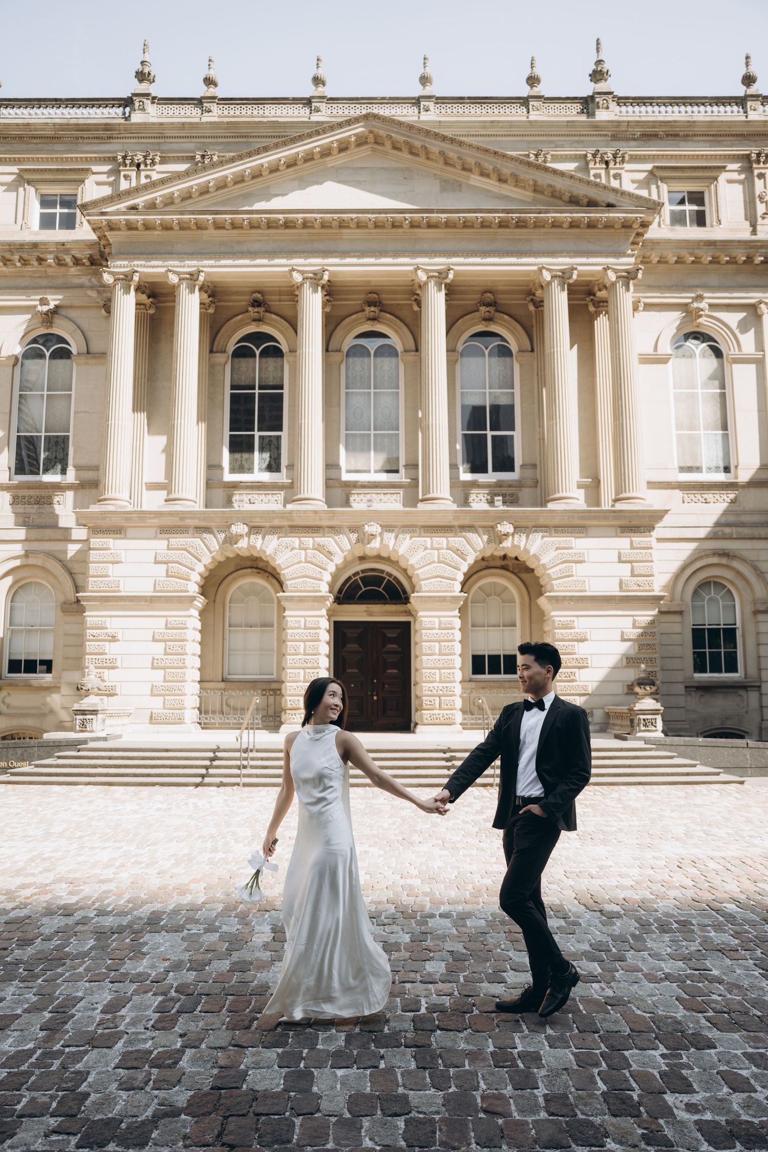 A couple in wedding attire dances on cobblestones in front of a grand neoclassical building with ornate columns and architecture.
