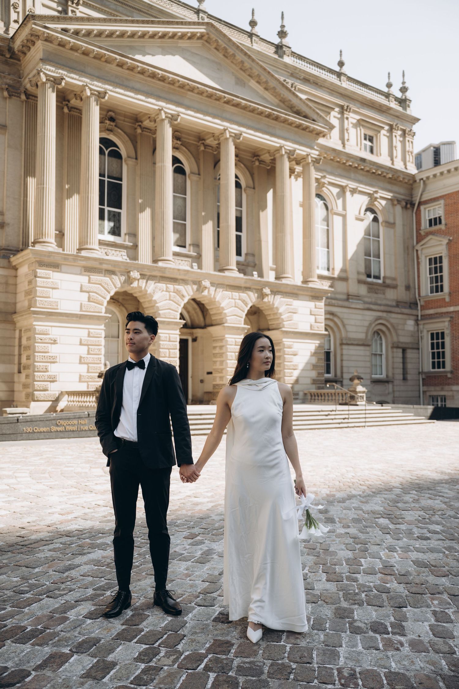 Elegant couple in wedding attire holds hands in front of grand historic building with classical columns and cobblestone courtyard.