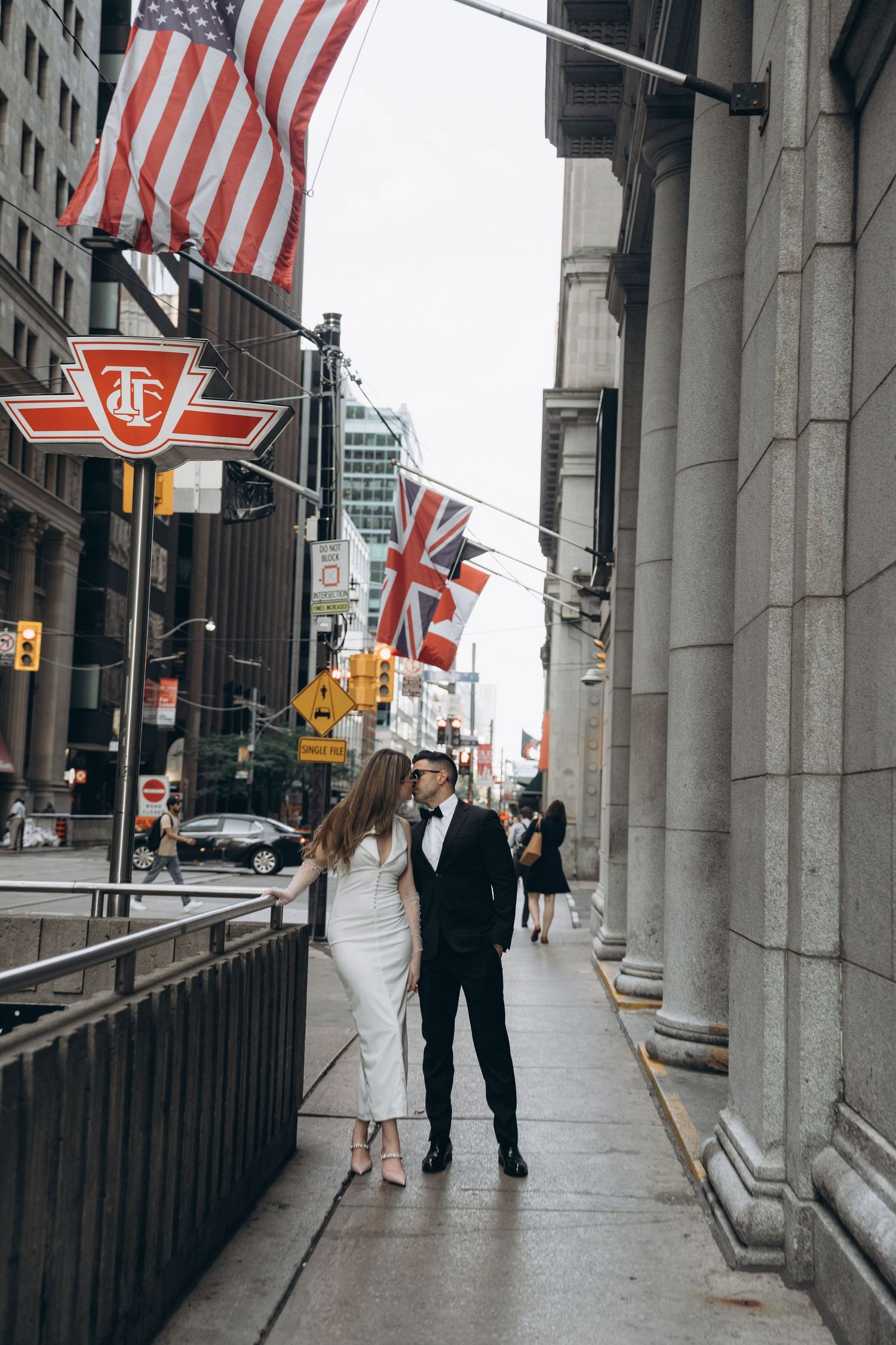 A couple walks down a city street with American and British flags hanging above near stone columns and the TTC logo.