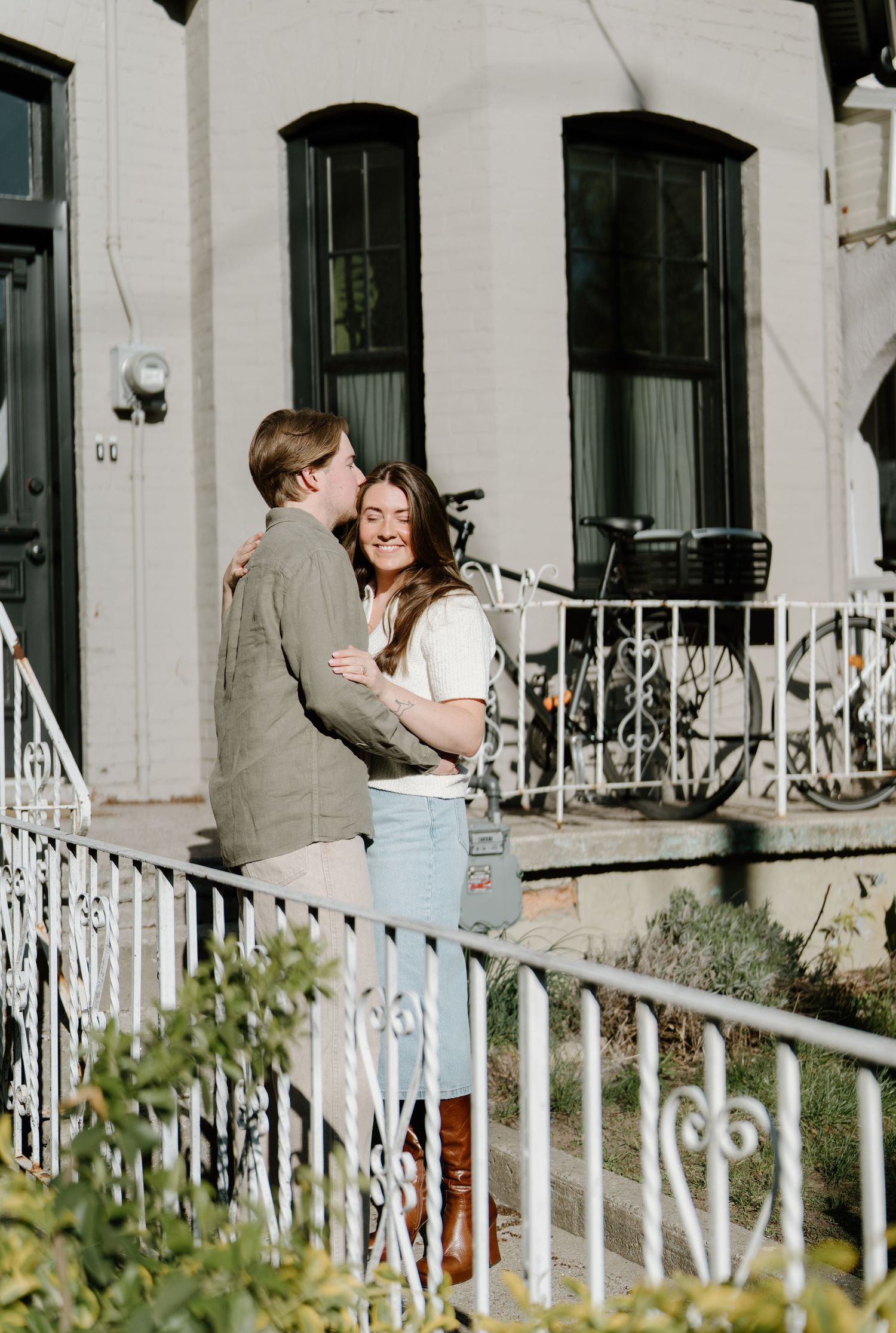 A couple embraces on white ornate porch steps of a classic townhouse with bikes visible in the background.