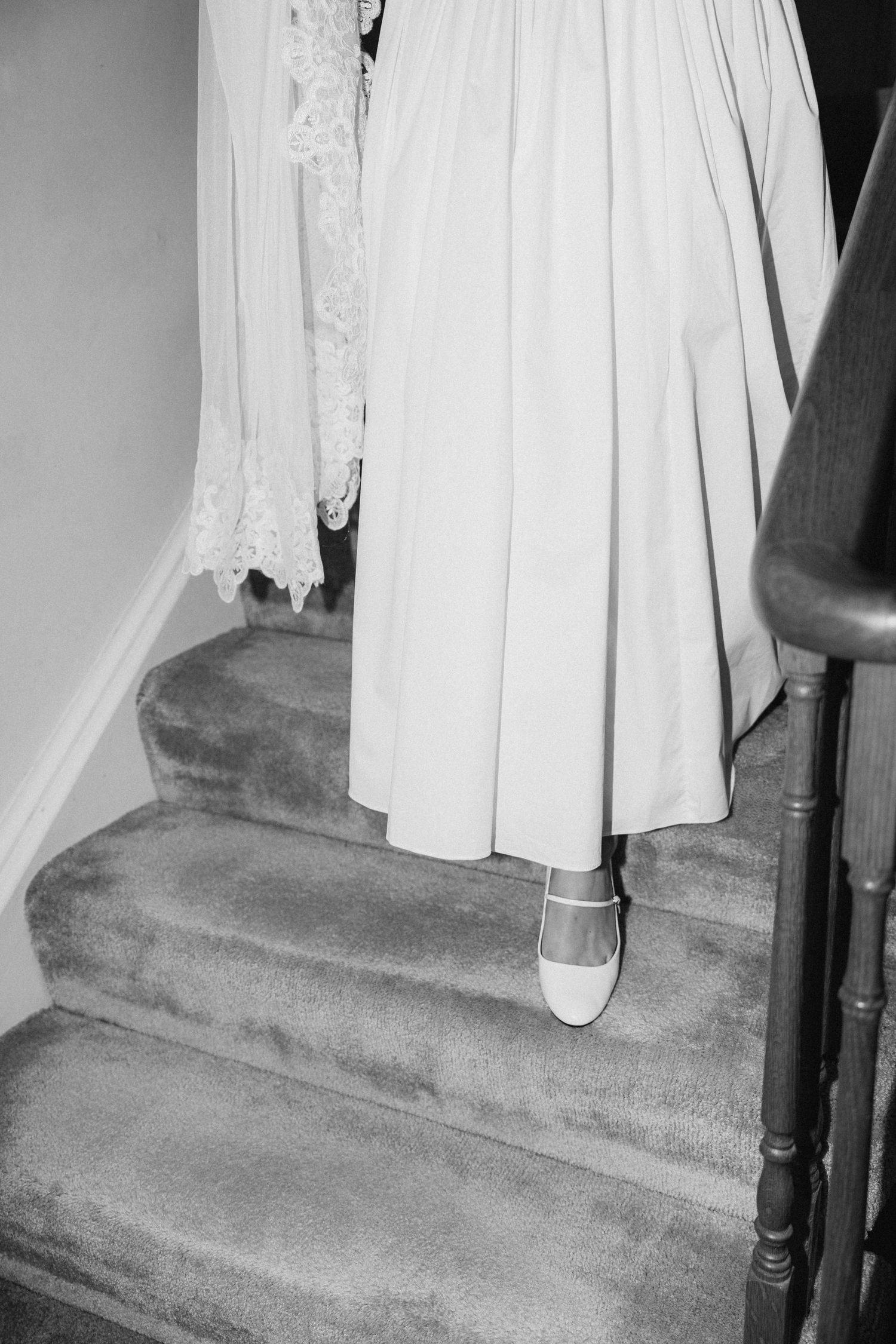 The hem of a white wedding dress shown against carpeted stairs with dark wooden banisters.