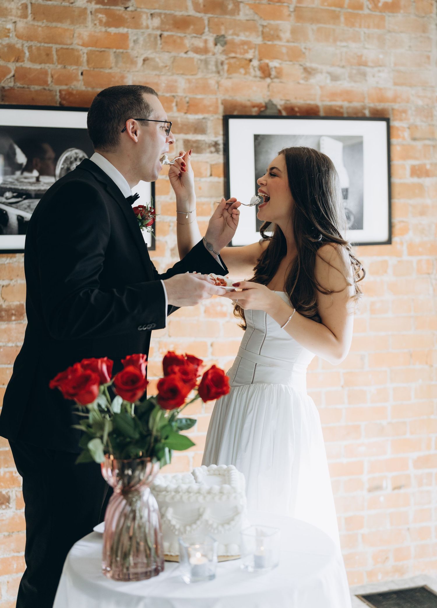 A couple feeds each other cake during their wedding celebration against an exposed brick wall with red roses in view.