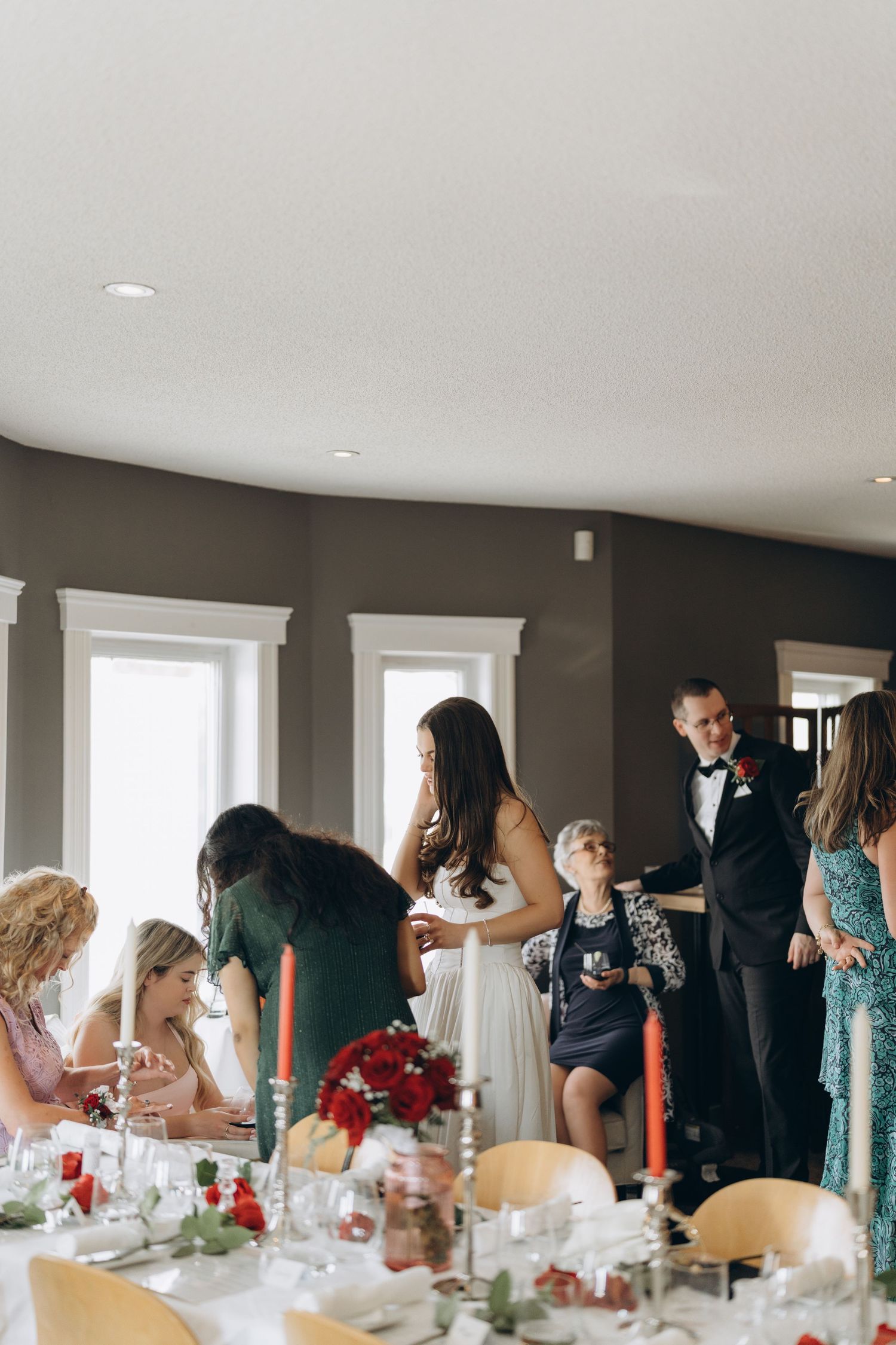Wedding guests mingling around elegantly set dining tables with white linens and red floral centerpieces.