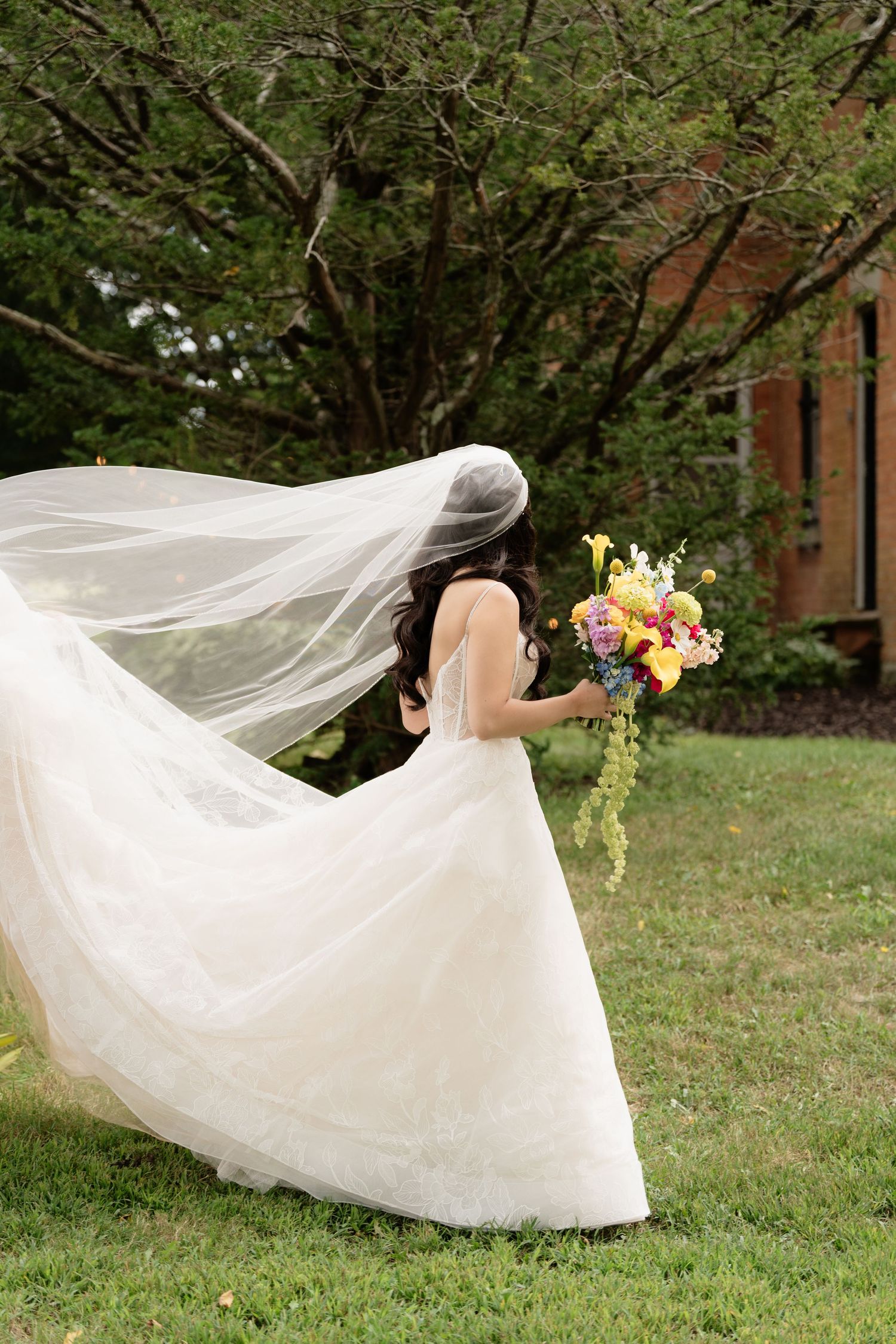 A bride in a flowing white wedding dress holds a colorful bouquet as her veil catches the breeze in a garden setting.