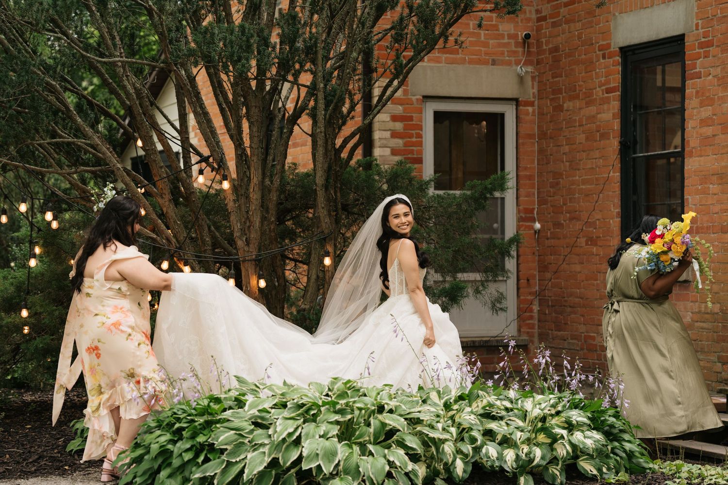 A bride in a flowing white wedding gown poses gracefully in front of a historic red brick building with greenery.