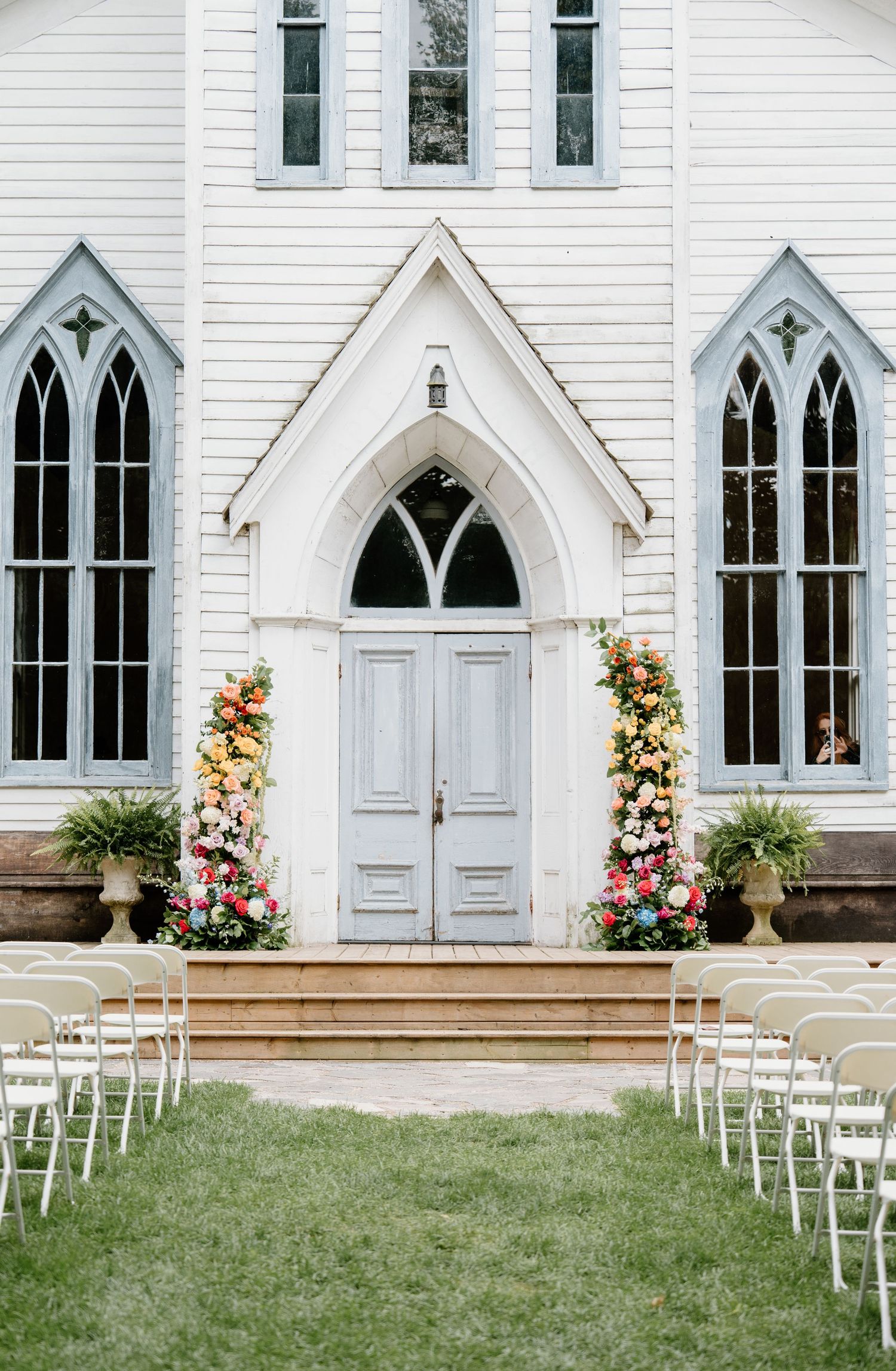 White chapel entrance with Gothic arched windows decorated with floral arrangements and white chairs set up for outdoor wedding ceremony.