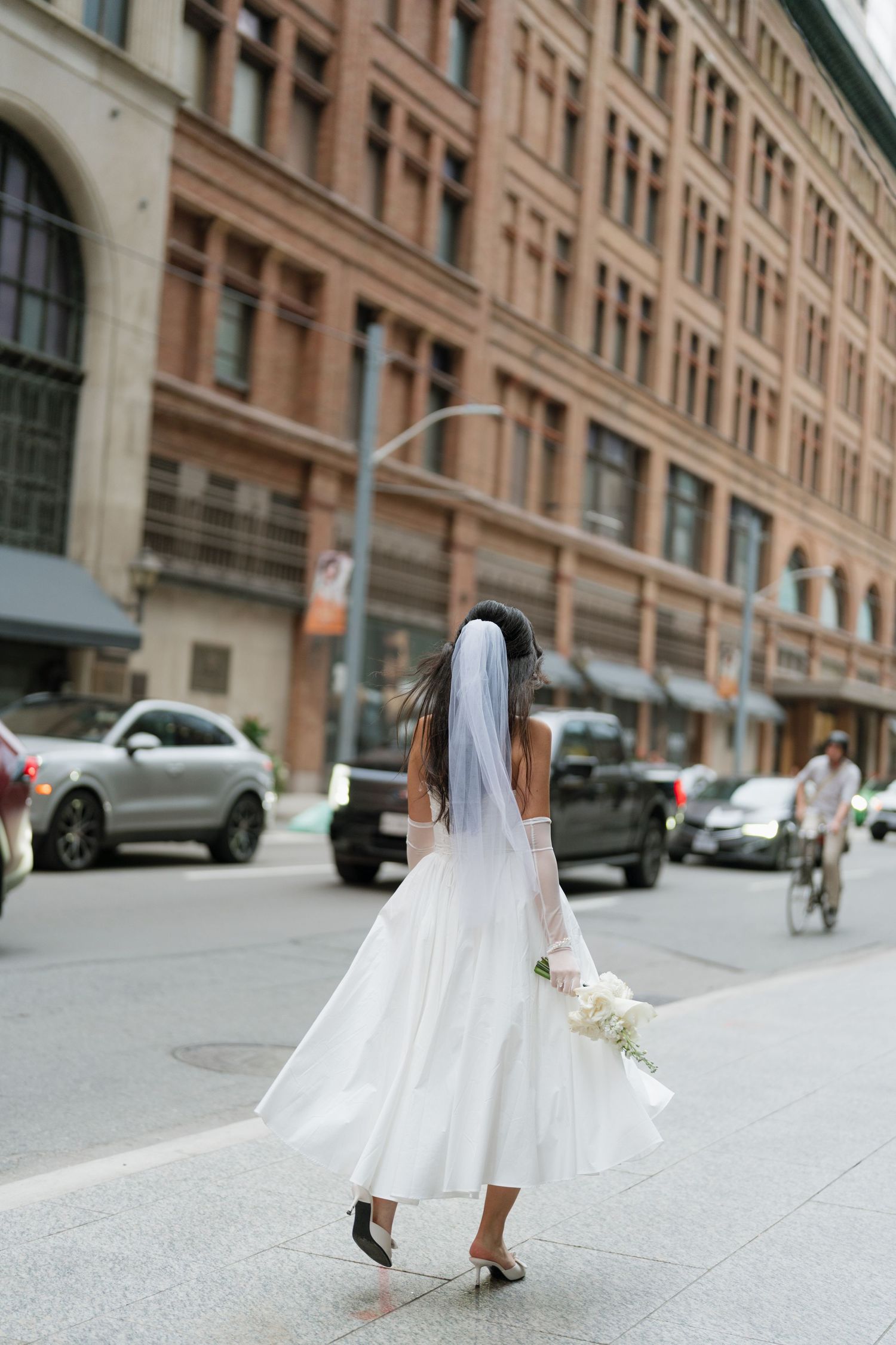 A bride in a flowing white dress and veil walks down a city street lined with historic brick buildings.