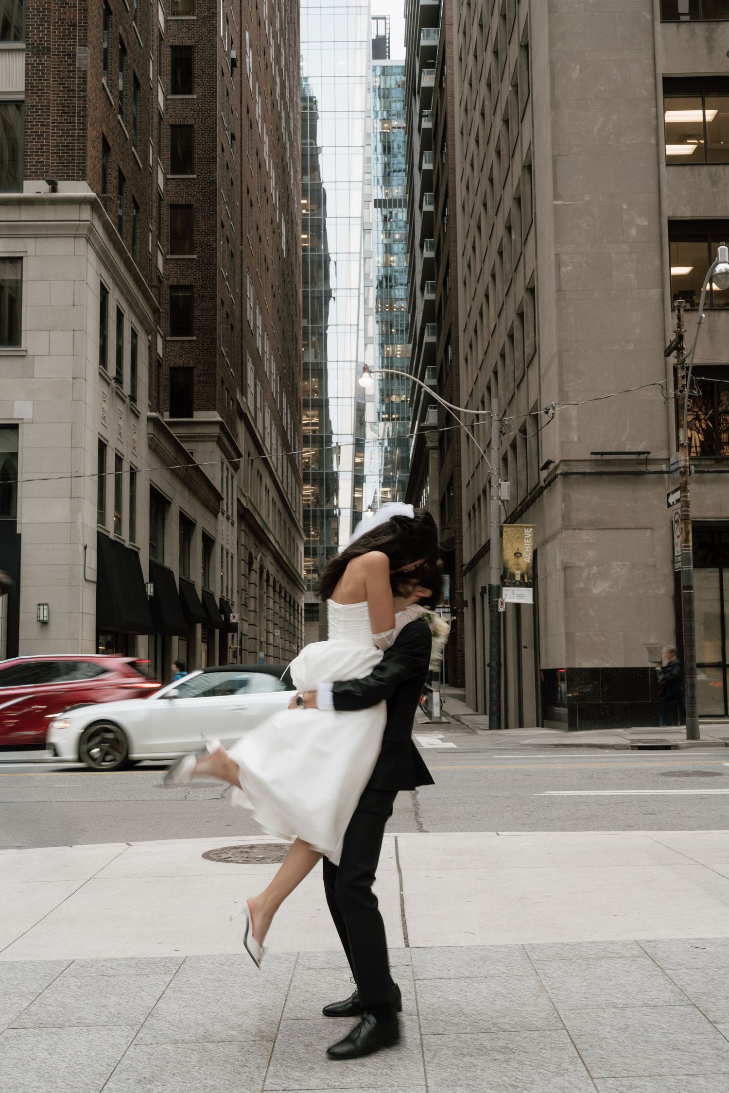 A couple in wedding attire performs a romantic dip pose in an urban setting with tall buildings in the background.