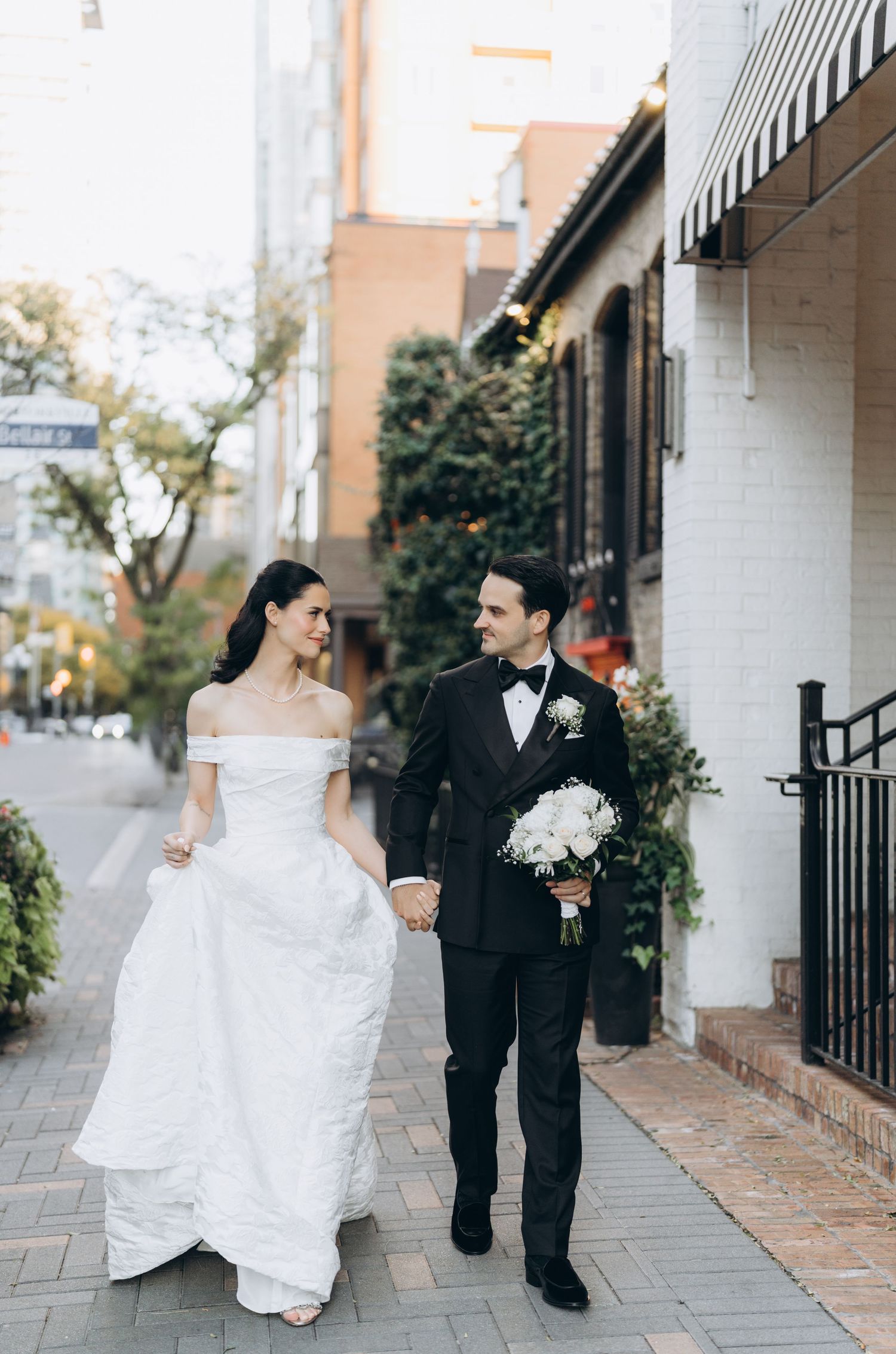 A couple in wedding attire walks down a charming city sidewalk with brick buildings and greenery in the background.