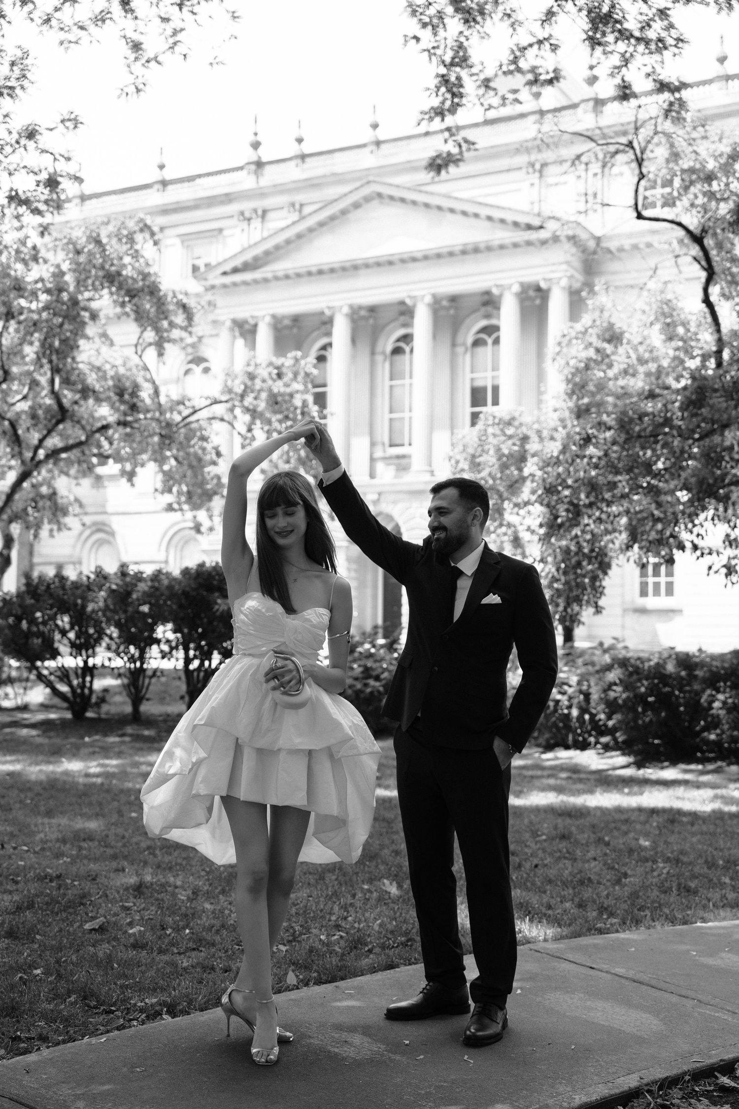 A couple in formal wedding attire performs a dancing sequence on a walkway outside an elegant white building.