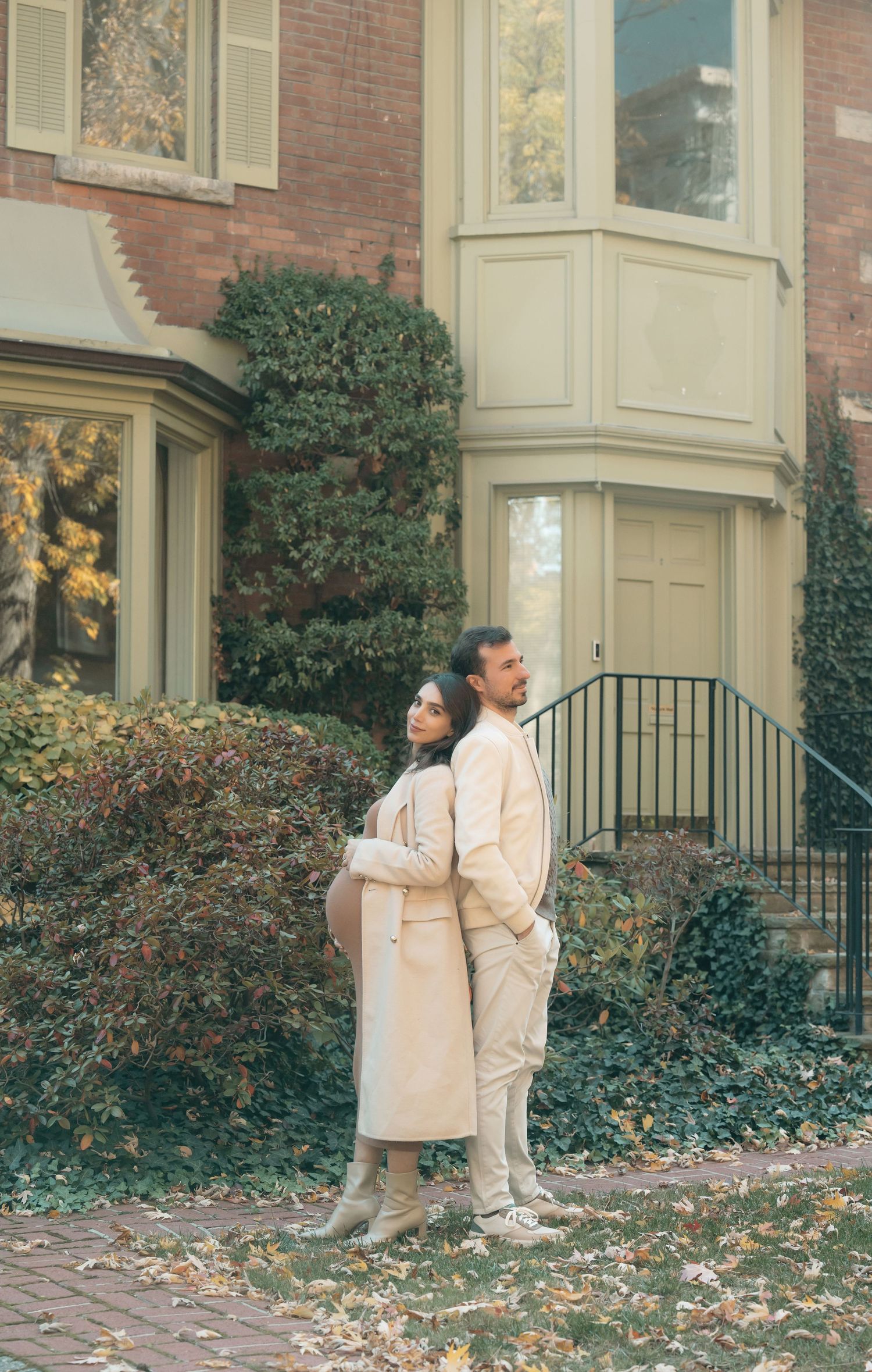 A couple in matching cream-colored coats stands back-to-back outside a brick building with autumn foliage.