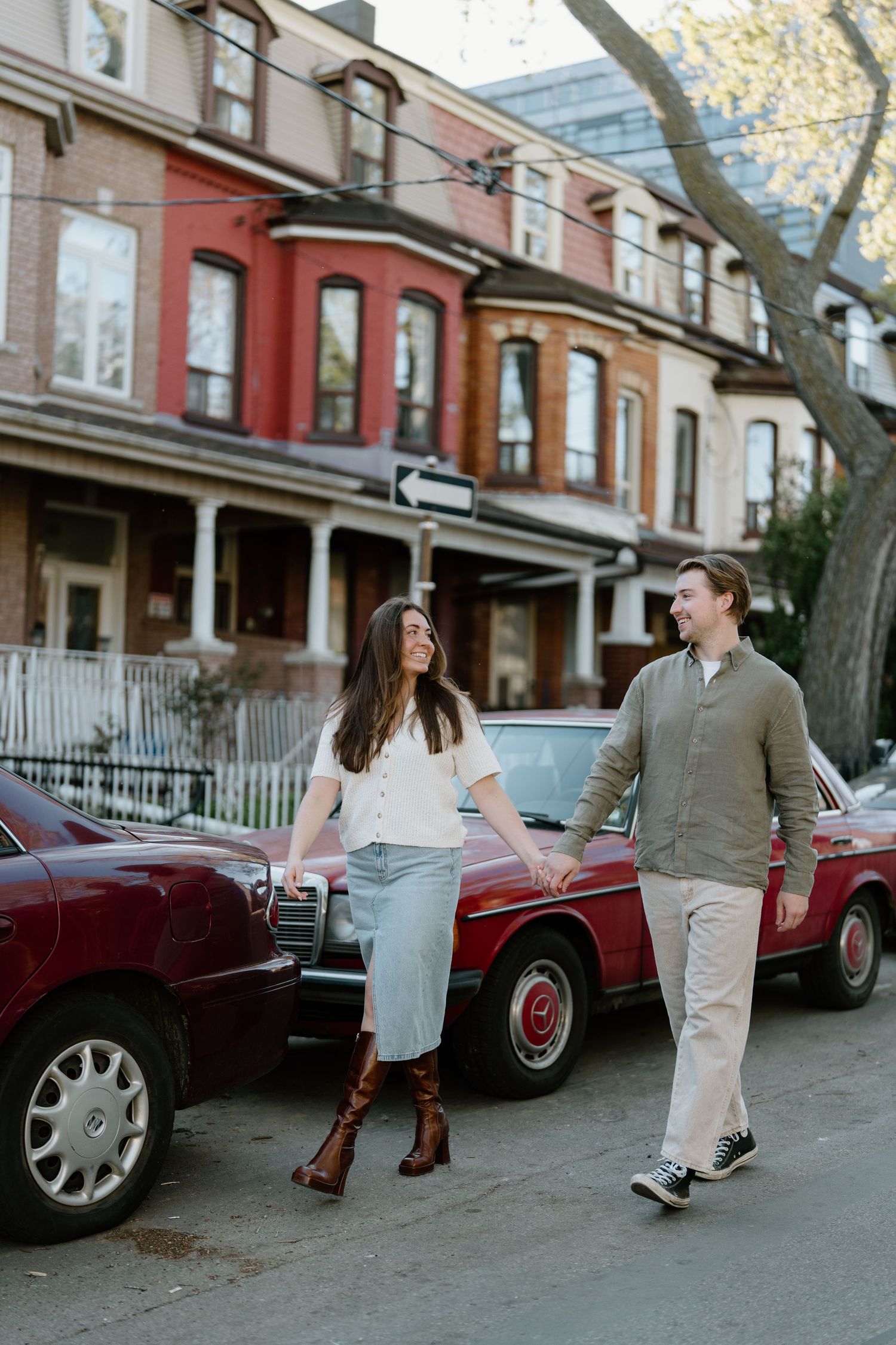 Couple walking and laughing together on a city street with vintage townhouses and a classic red car in the background.