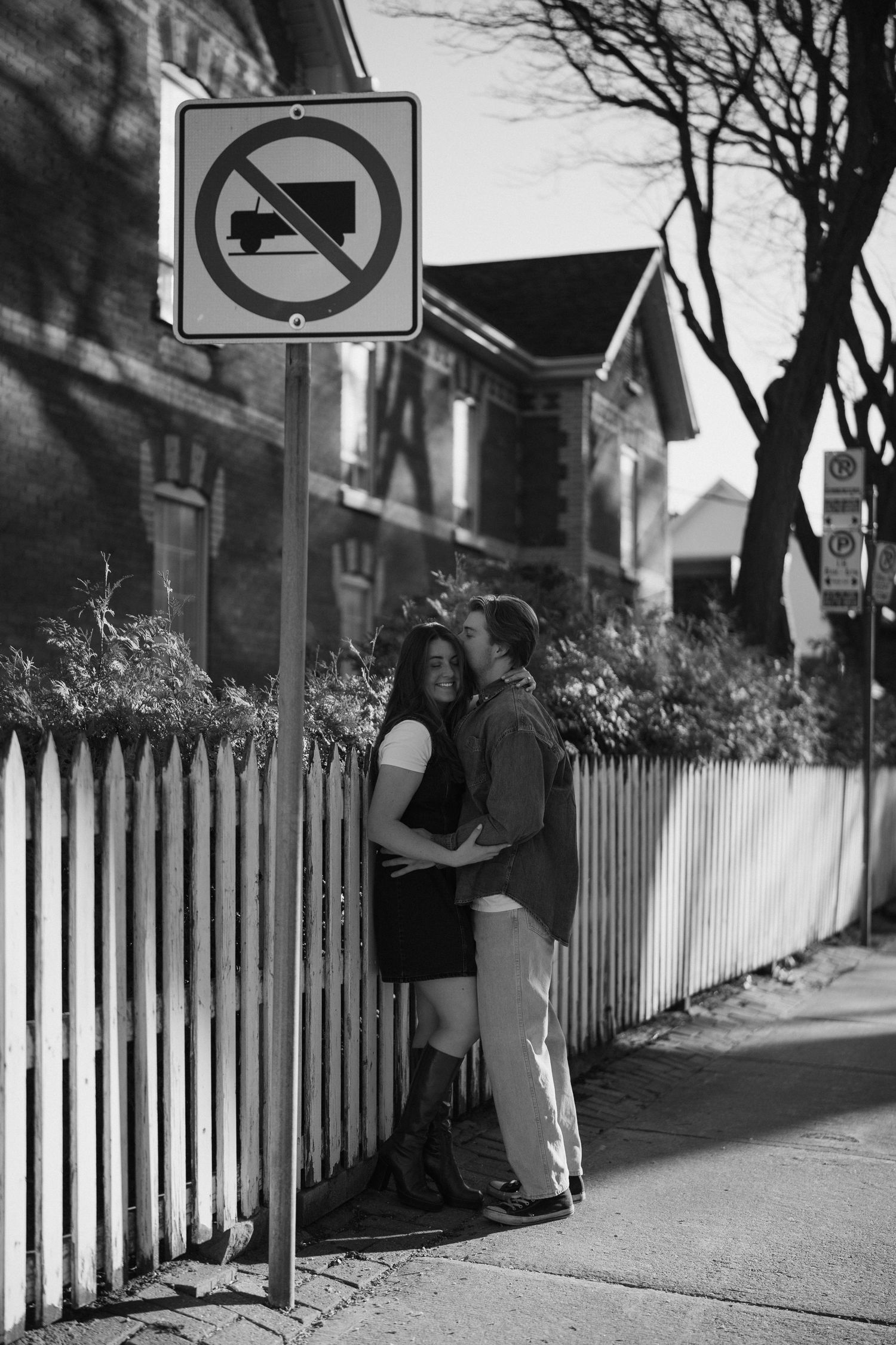 A couple embraces beside a white picket fence and no trucks sign on a residential street.