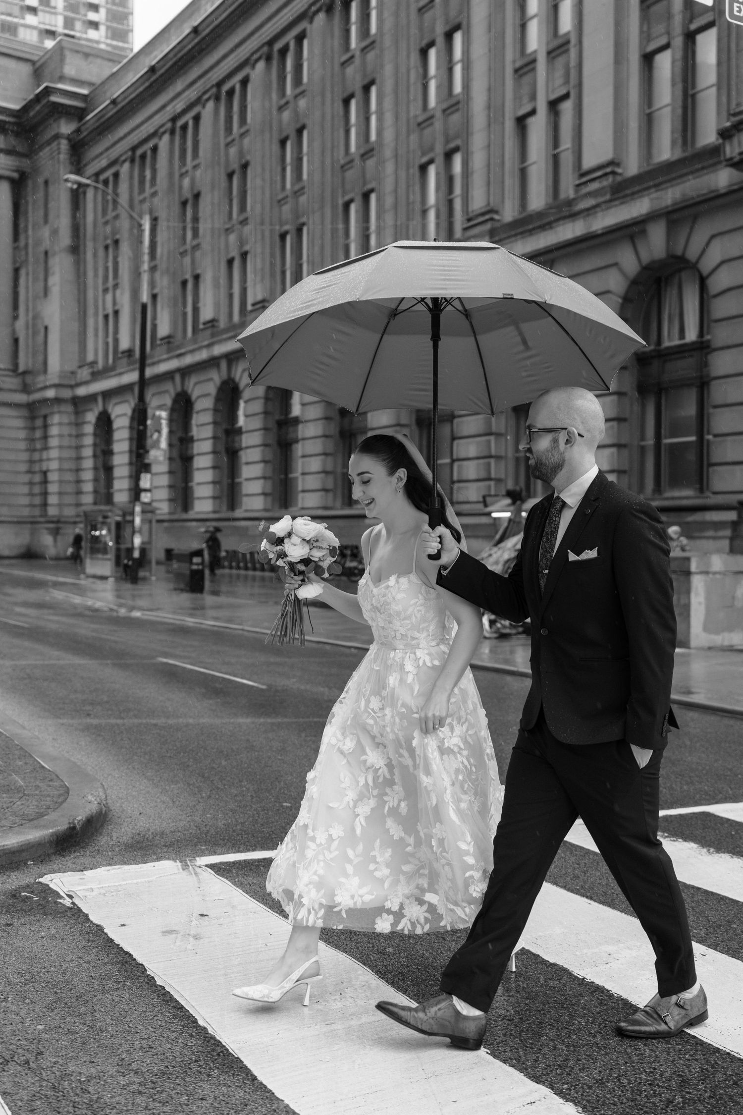 Two people walk under a white umbrella in the rain outside a historic stone building, one in a formal dress carrying flowers.