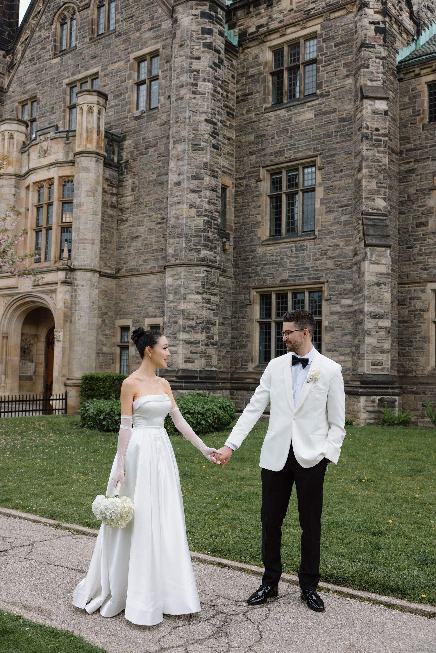 Newlywed couple holding hands in front of a grand stone castle building with Gothic architecture on their wedding day.