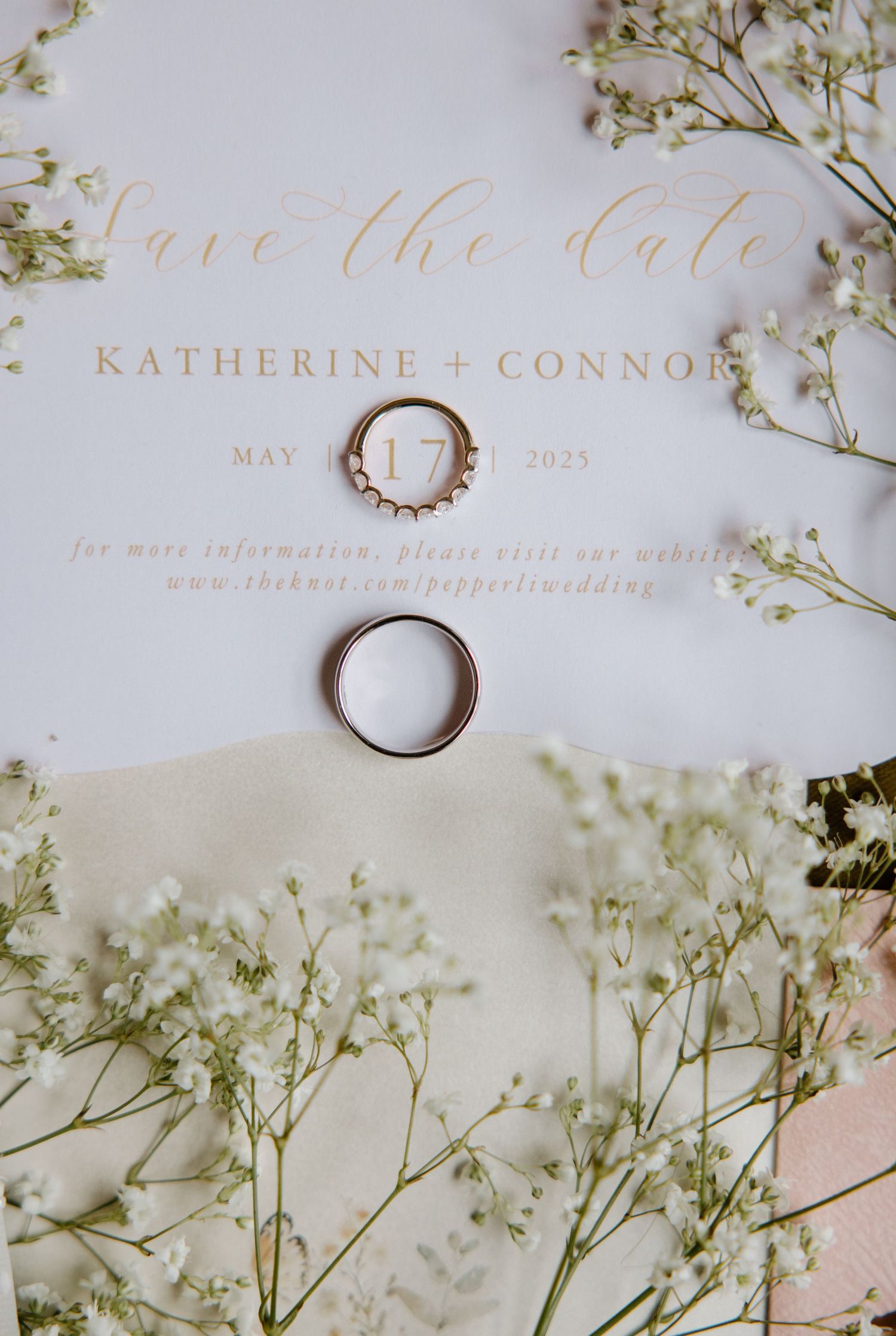 Wedding save the date invitation with gold calligraphy text, wedding rings, and delicate baby's breath flowers on white paper.