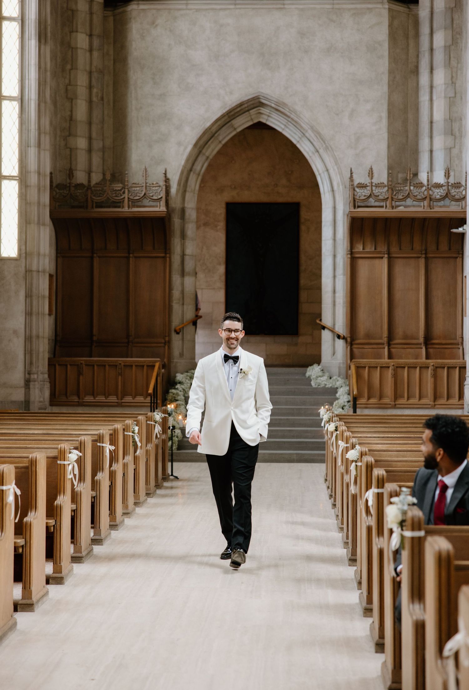 A groom walks down the center aisle of a grand church with wooden pews and a Gothic stone archway.