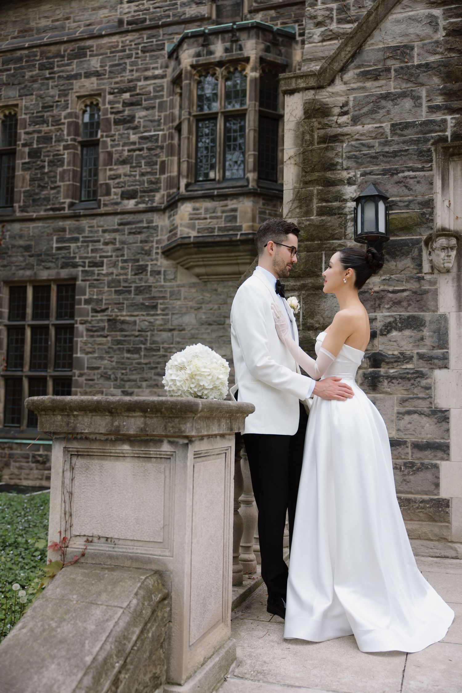 A couple in white wedding attire stands together against a stone building facade in an elegant pose.