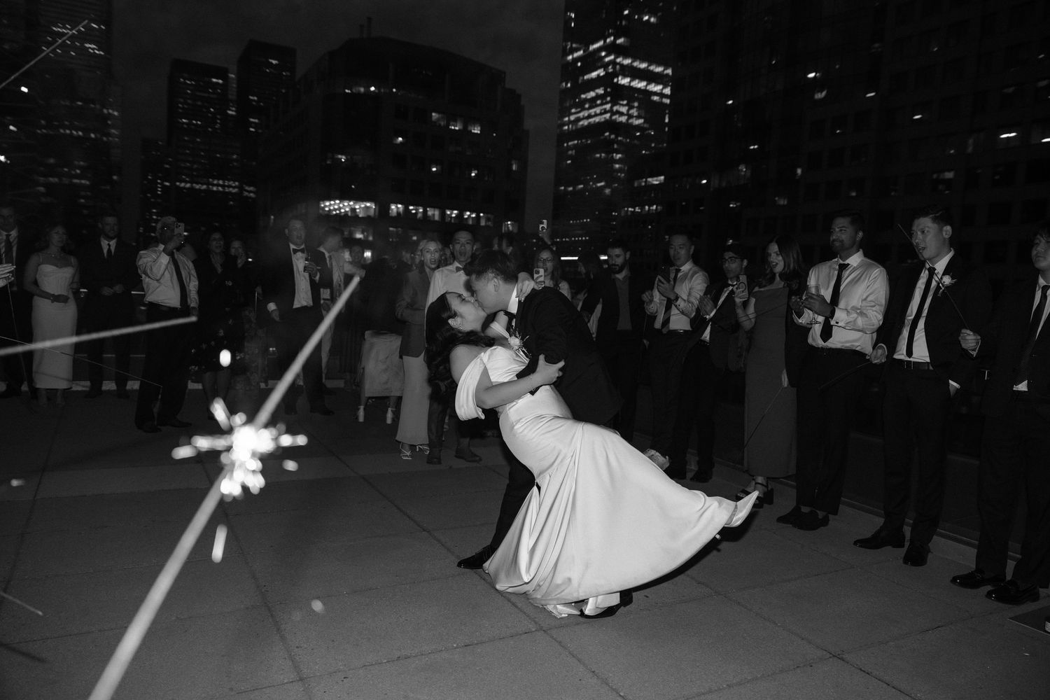 A couple performs a romantic dip dance move at a nighttime outdoor reception while sparklers glow in the foreground.