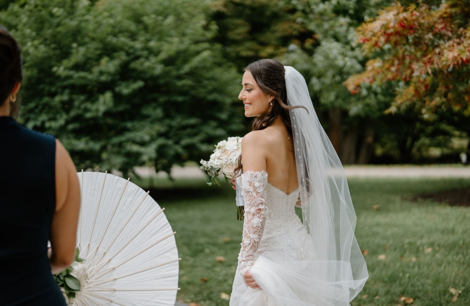 A wedding party poses outdoors with a white parasol against a green garden backdrop in autumn.