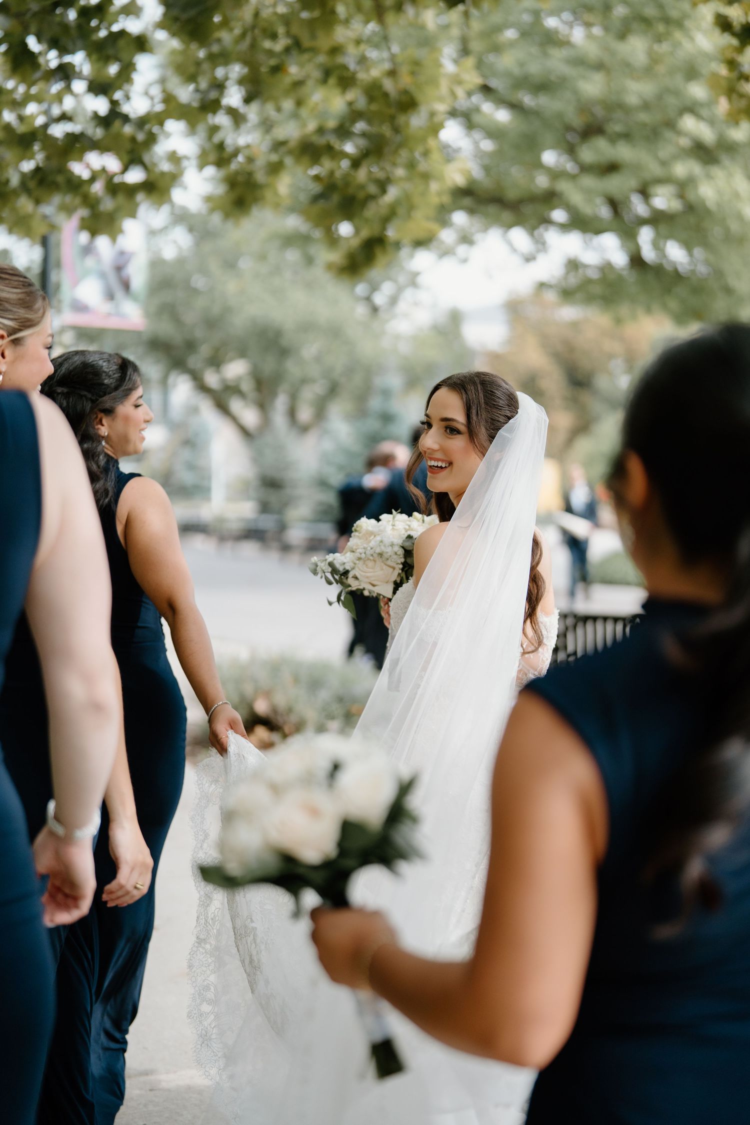 A bride in a white wedding gown and veil smiles while holding a bouquet, surrounded by bridesmaids in navy dresses.