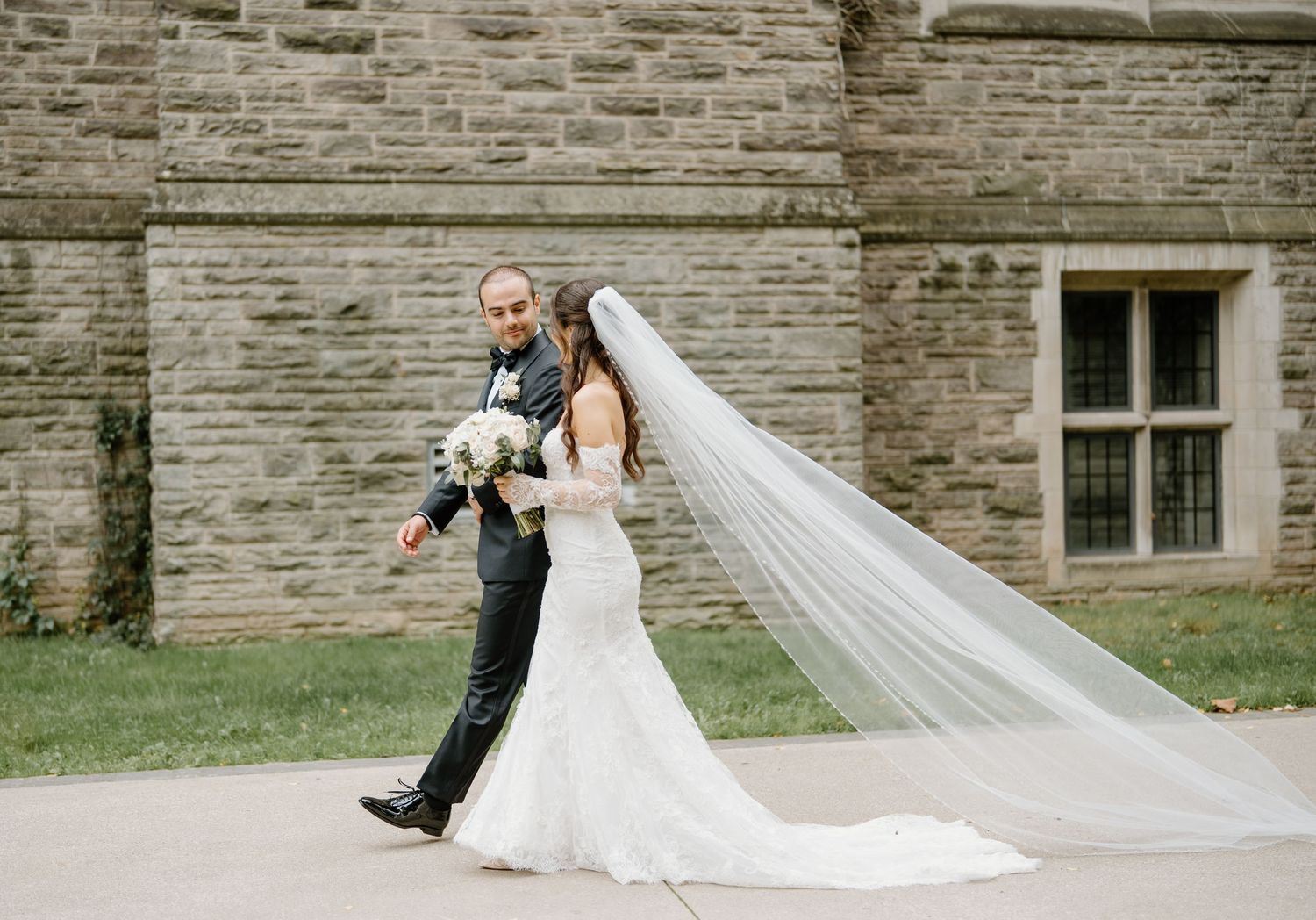 A bride and groom pose for a creative wedding photo series against a stone wall, with a flowing white veil trailing behind.