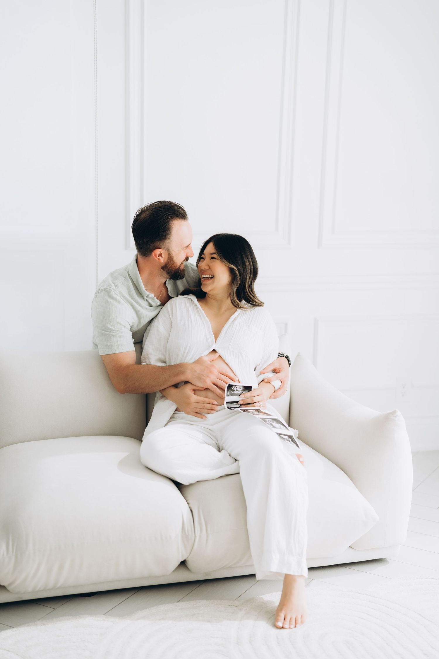 A couple dressed in white shares a tender moment while sitting together on a light-colored couch.