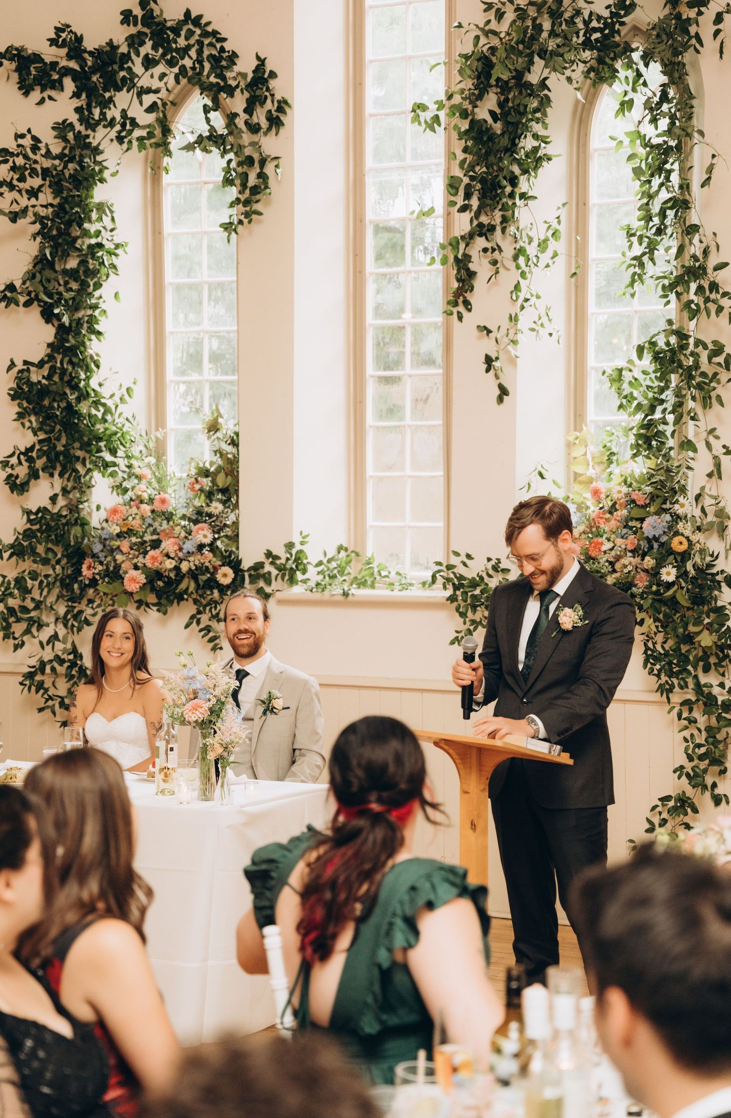 Wedding reception in an elegant venue decorated with hanging greenery and flowers during speeches.