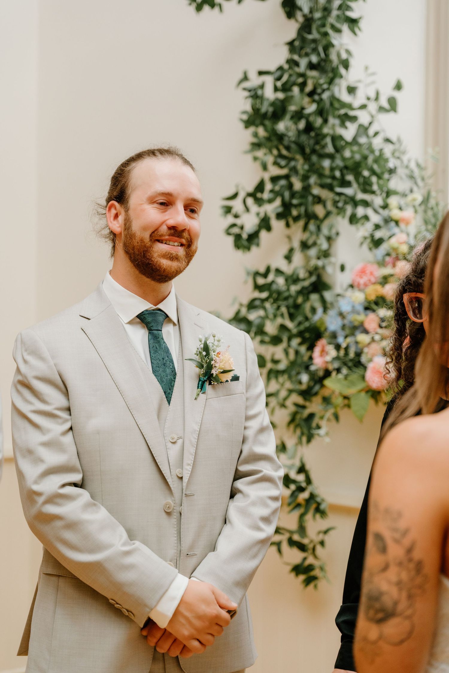 Groom in light beige suit with blue tie smiles during wedding ceremony against floral backdrop.