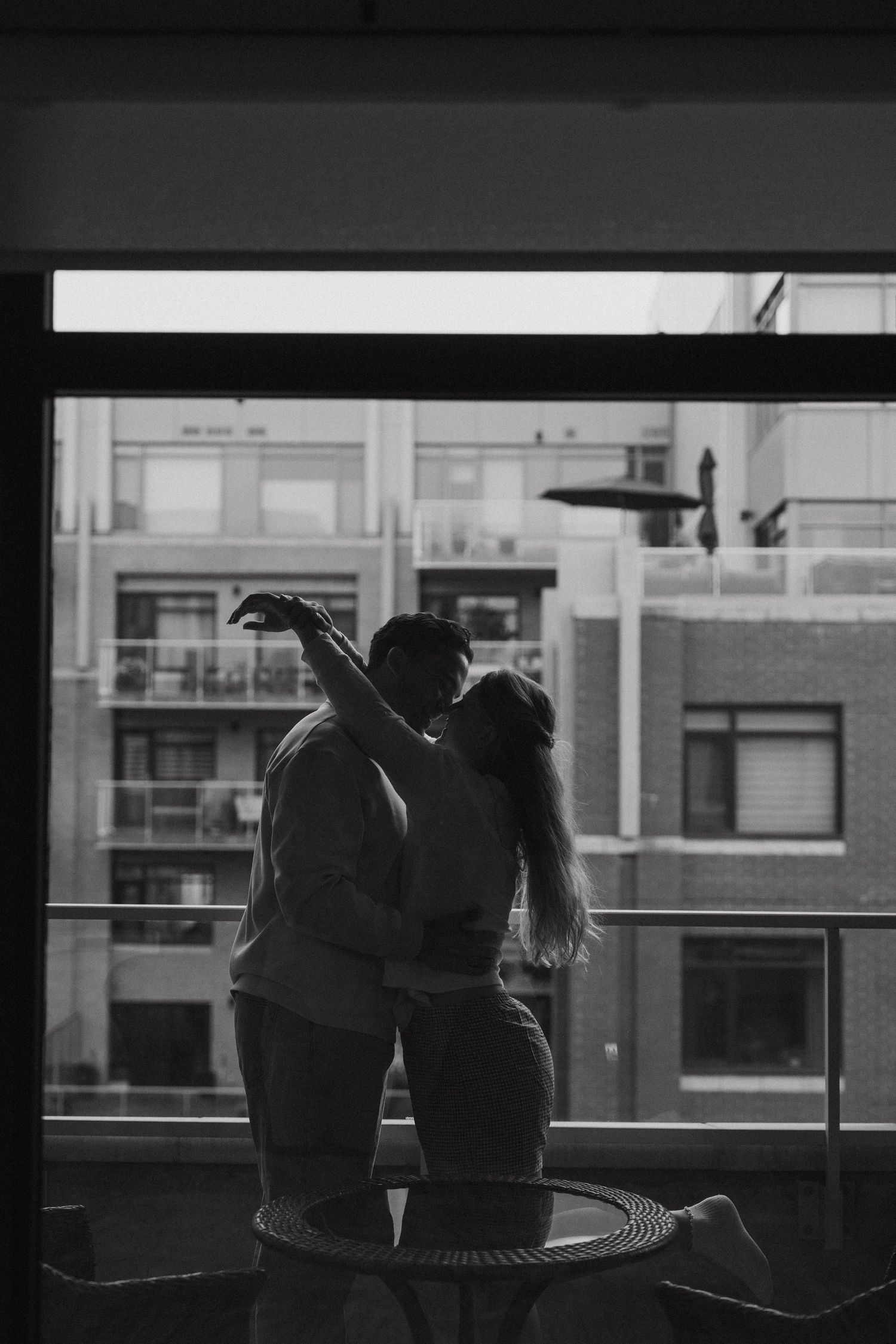 A silhouetted couple shares an intimate moment on a balcony against an urban backdrop in black and white.