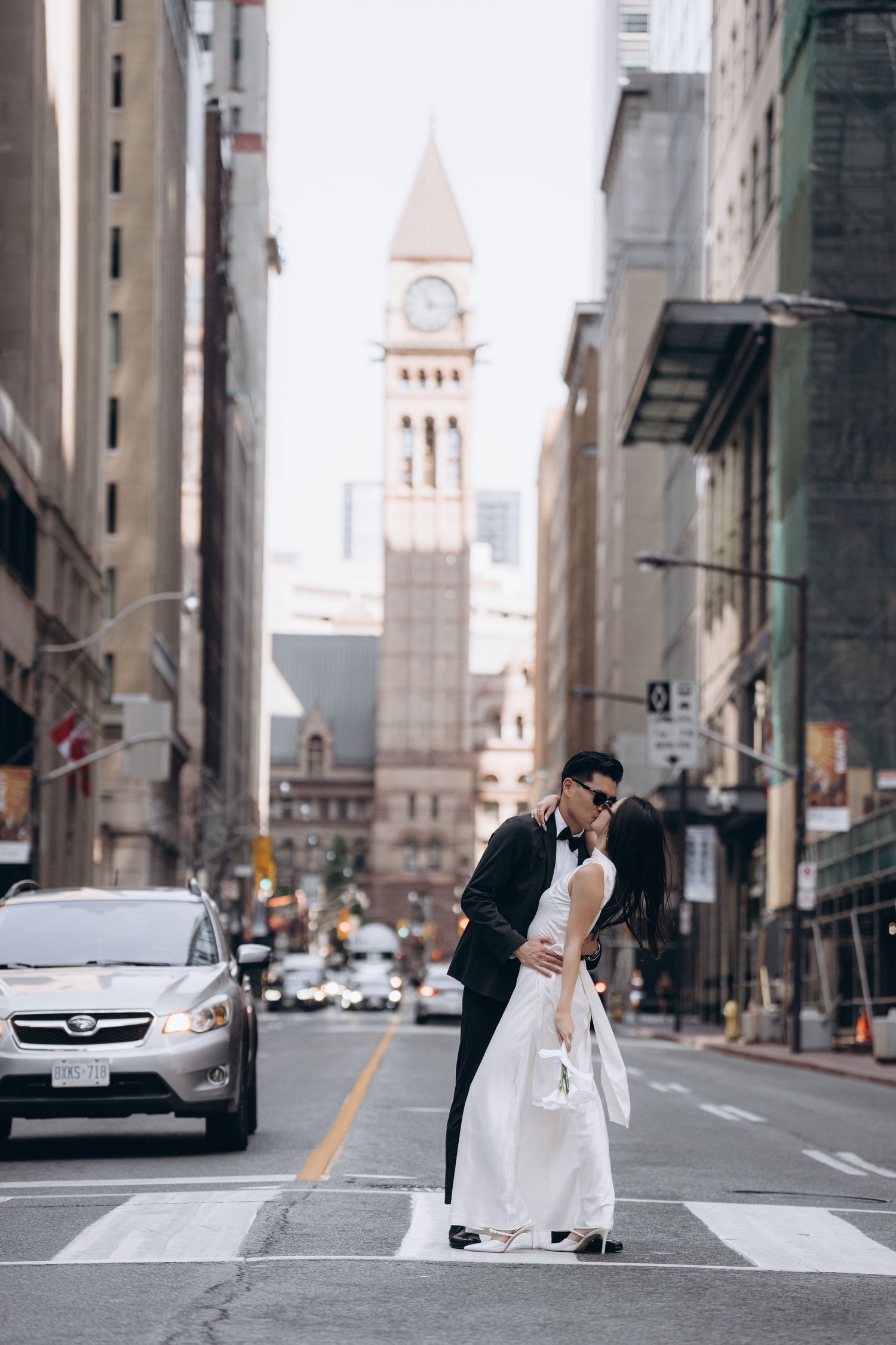 Romantic couple shares a kiss on a city crosswalk with a historic clock tower and church spire visible in the background.